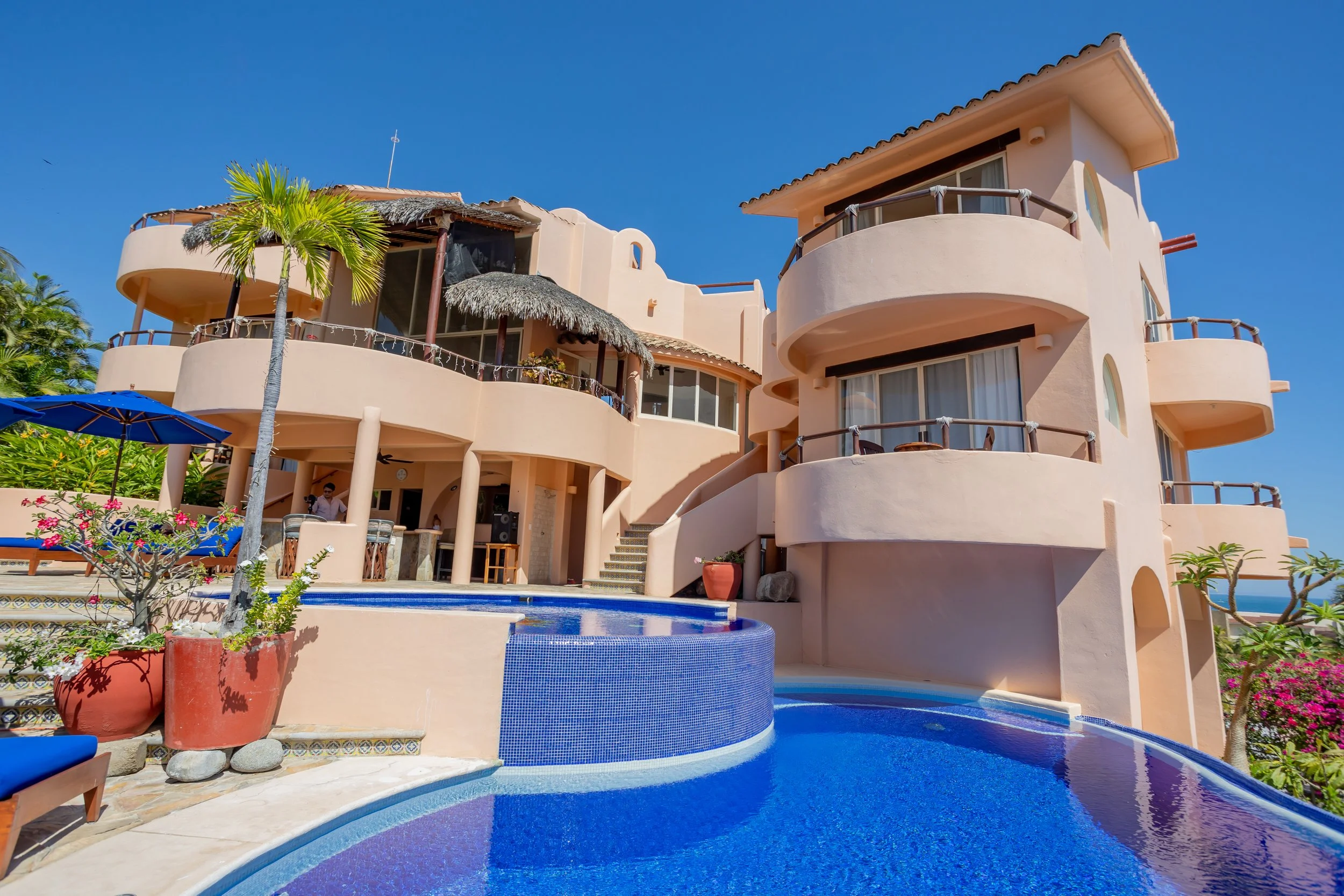 A multi-story peach-colored building with curved balconies, large glass windows, and a thatched roof section, set against a bright blue sky. There is a swimming pool in the foreground with blue tiles and surrounding potted plants and umbrellas.