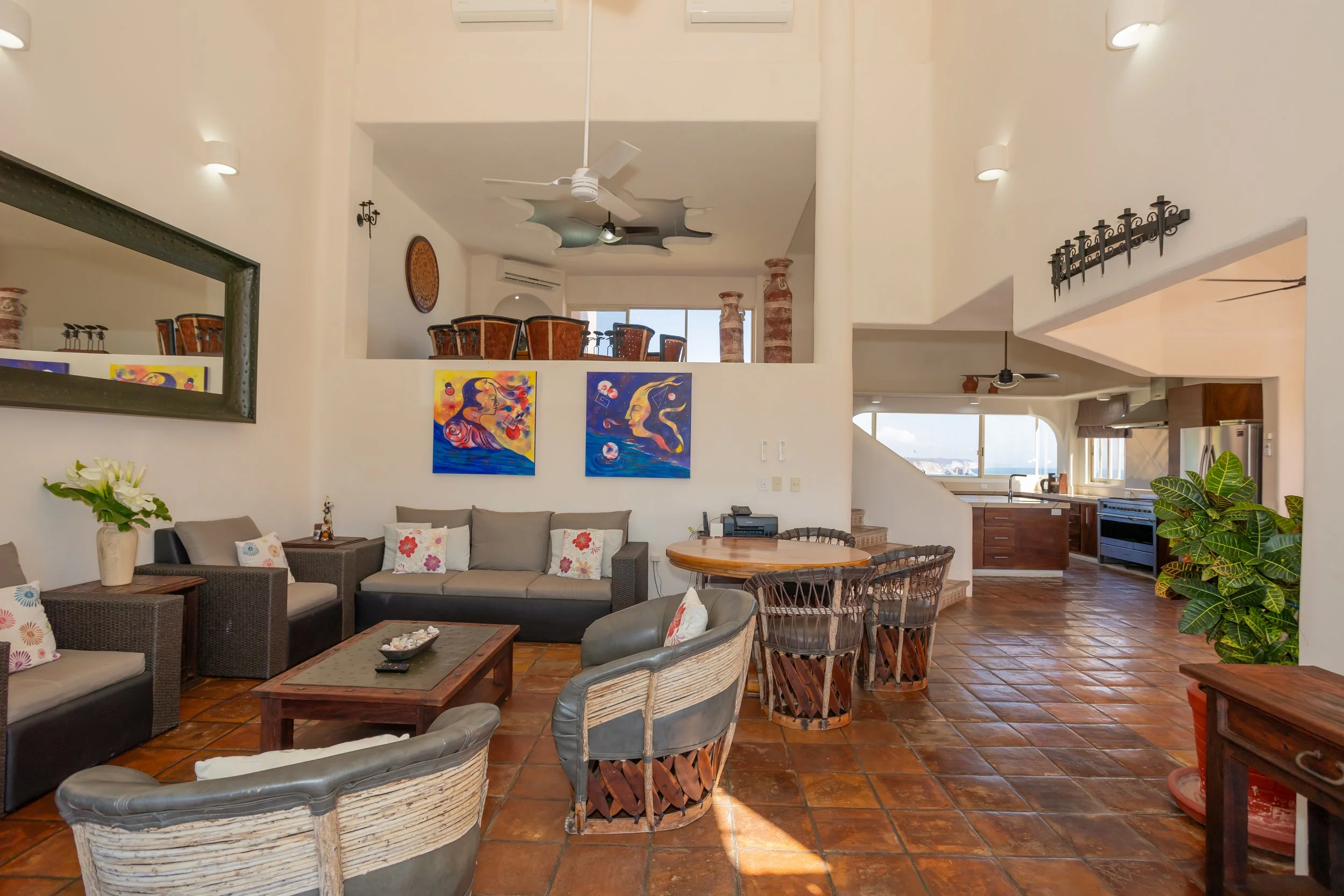 Interior of a living room with a beige sofa, wicker chairs, colorful pop art style paintings on the wall, a large mirror, and a view of the kitchen with wooden cabinets and stainless steel appliances.