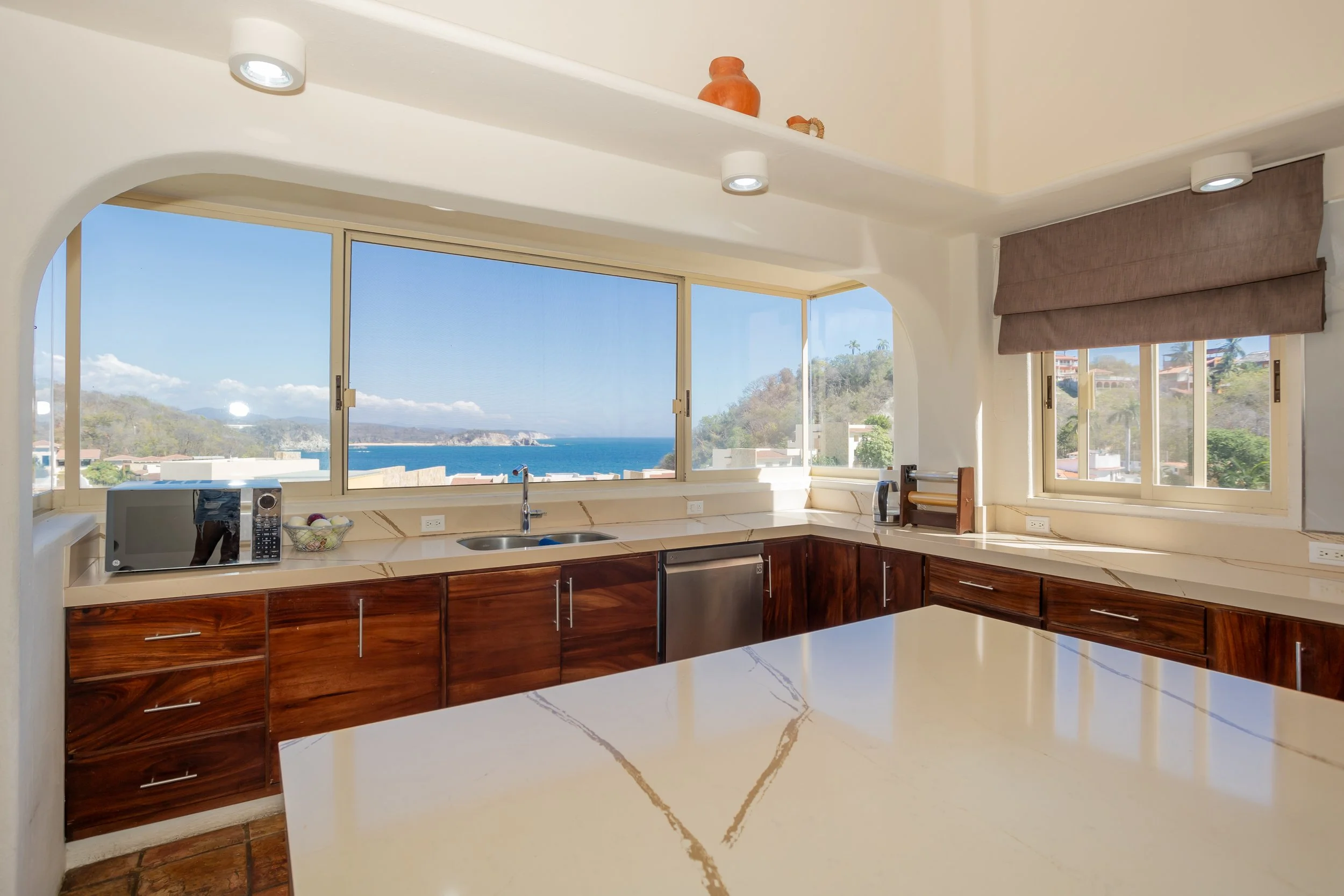A kitchen with large windows showing a view of hills, water, and blue sky. The kitchen has brown wooden cabinets, a white countertop, a microwave, and a sink.