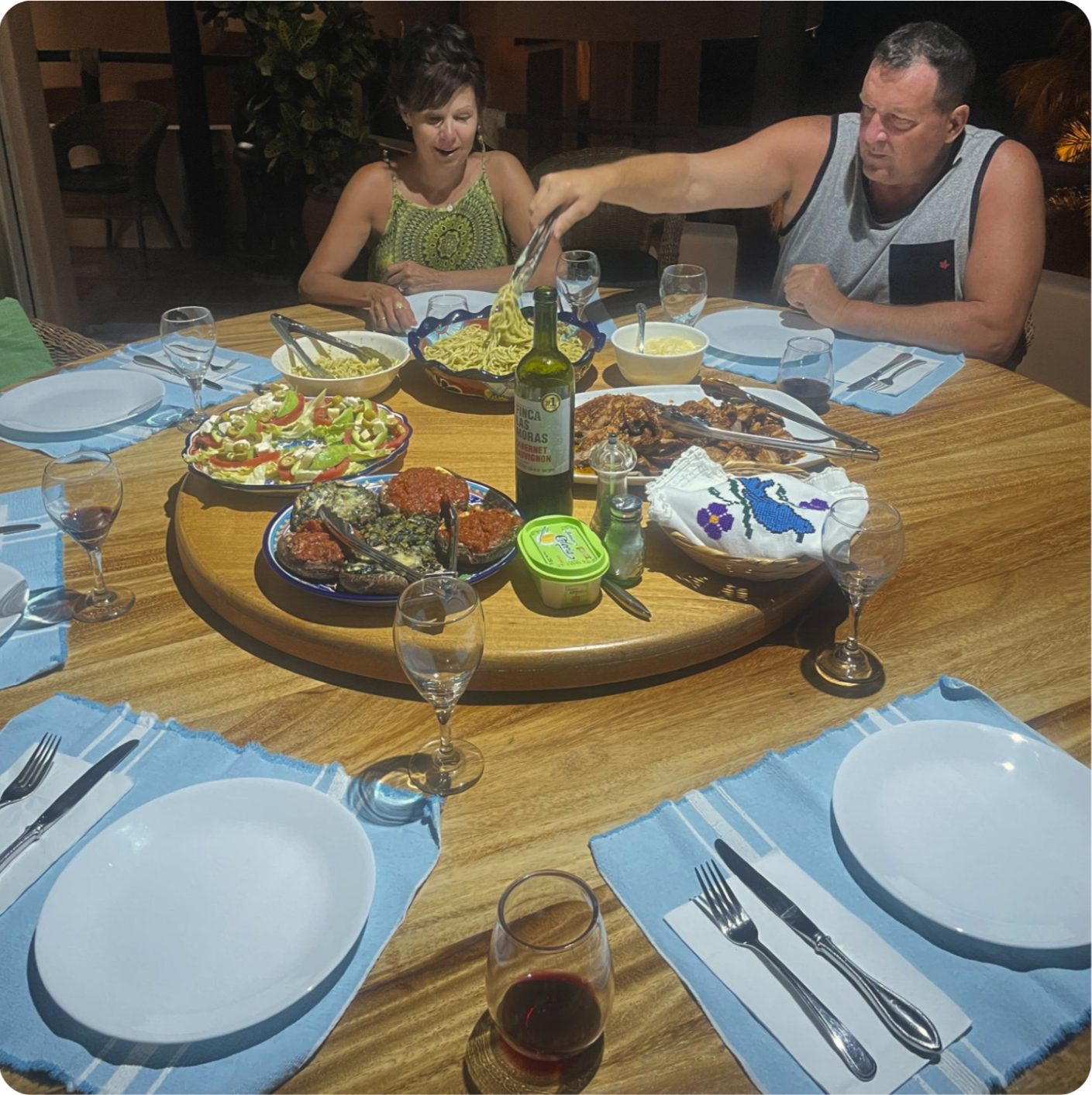 A family dinner with a round wooden table set with white plates, blue placemats, silverware, and glasses, featuring various Italian dishes including pasta, an antipasto platter, a bottle of wine, and a bowl of soup. Two people are actively serving or eating, one woman reacting with surprise, and a man reaching for the food.