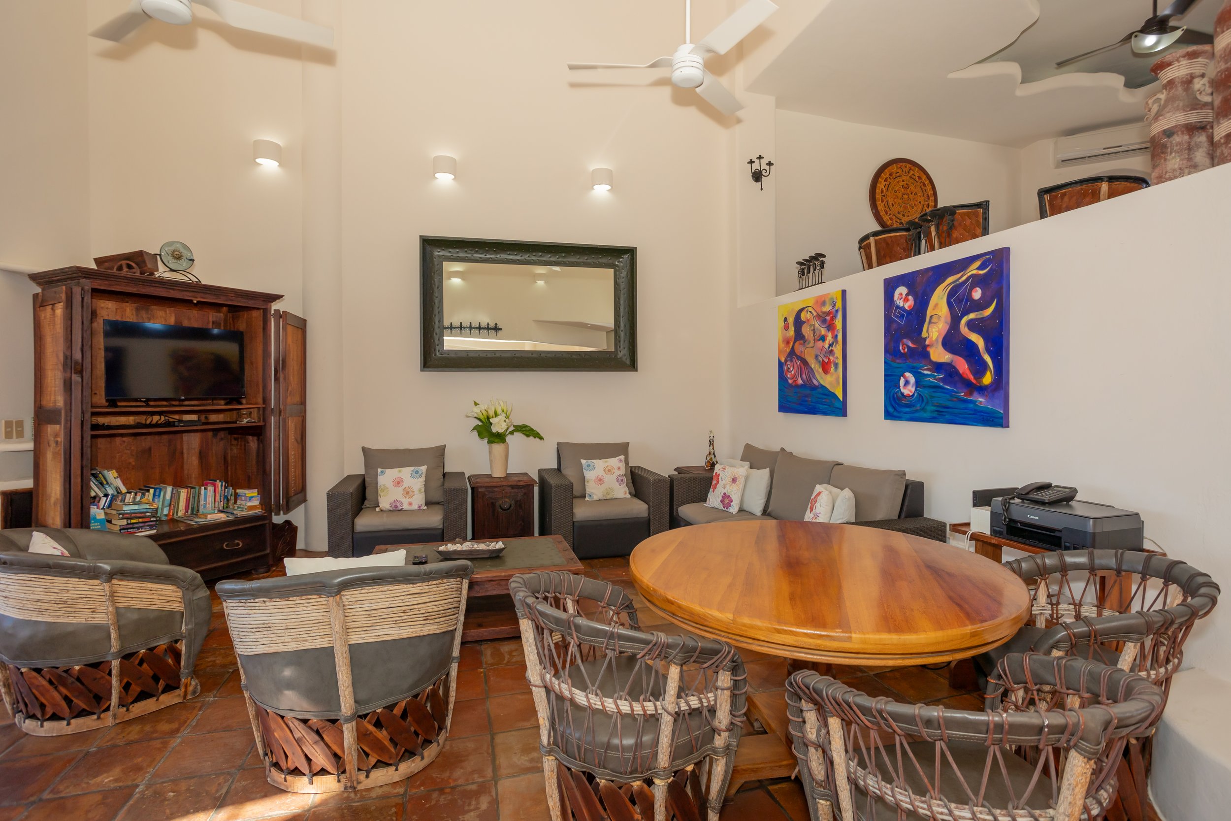 Living room with beige walls, ceiling fans, wall-mounted lights, a large mirror, colorful artwork, wooden furniture, a flat-screen TV, books, plants, and a round dining table with wicker chairs.
