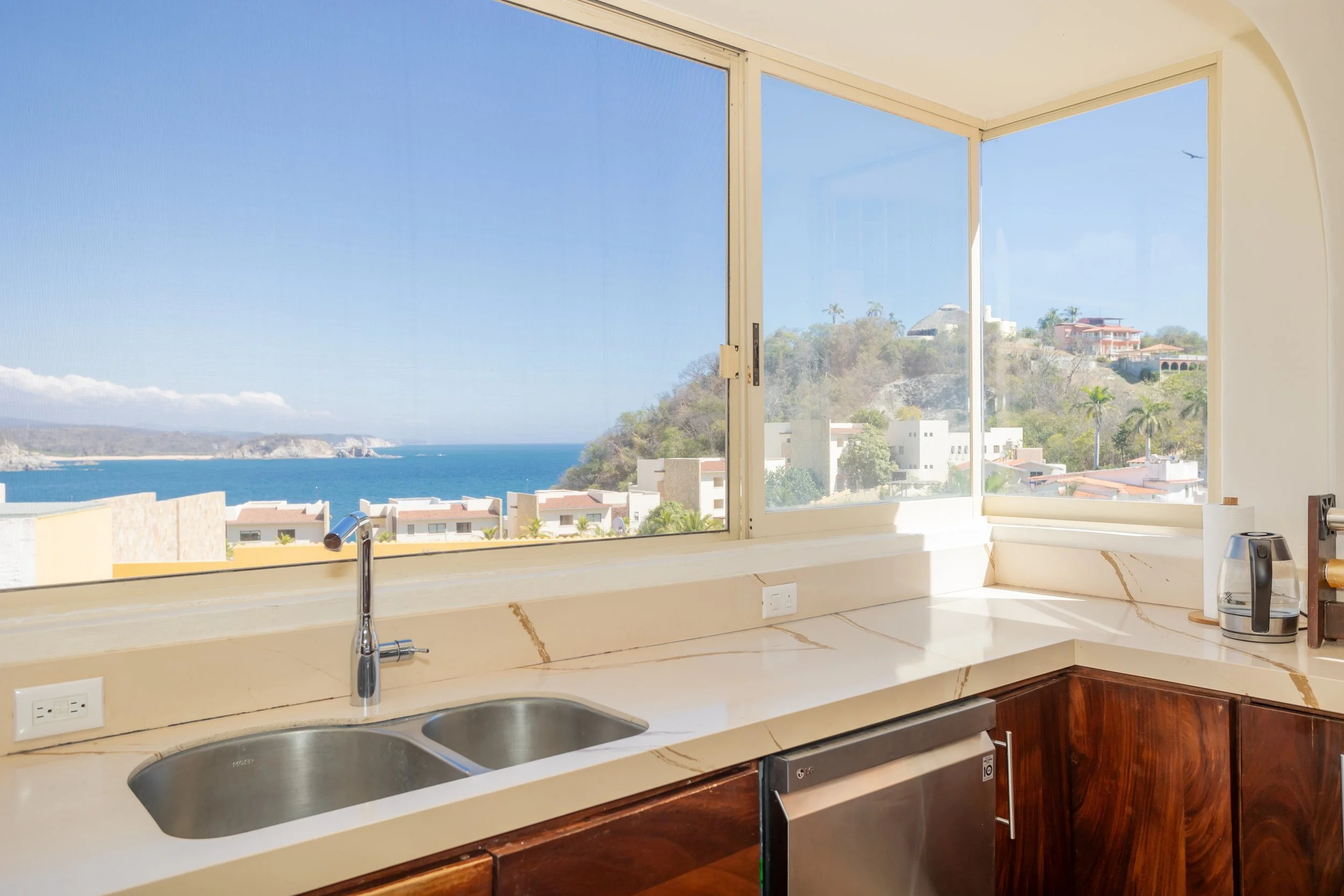 A kitchen with large corner window showing a view of the ocean, houses, and trees outside. The countertop has a double sink, a toaster, and an electric kettle.
