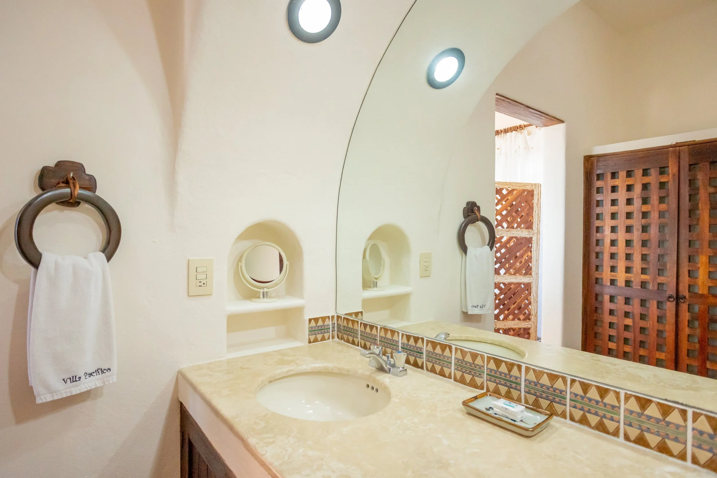 Bathroom countertop with beige marble surface, oval sink, silver faucet, decorative tile border, large mirror, two small round mirrors, wall-mounted towel ring with white towel labeled 'Villa Pacifico', wood cabinet door, window with sheer curtain, recessed ceiling lights.