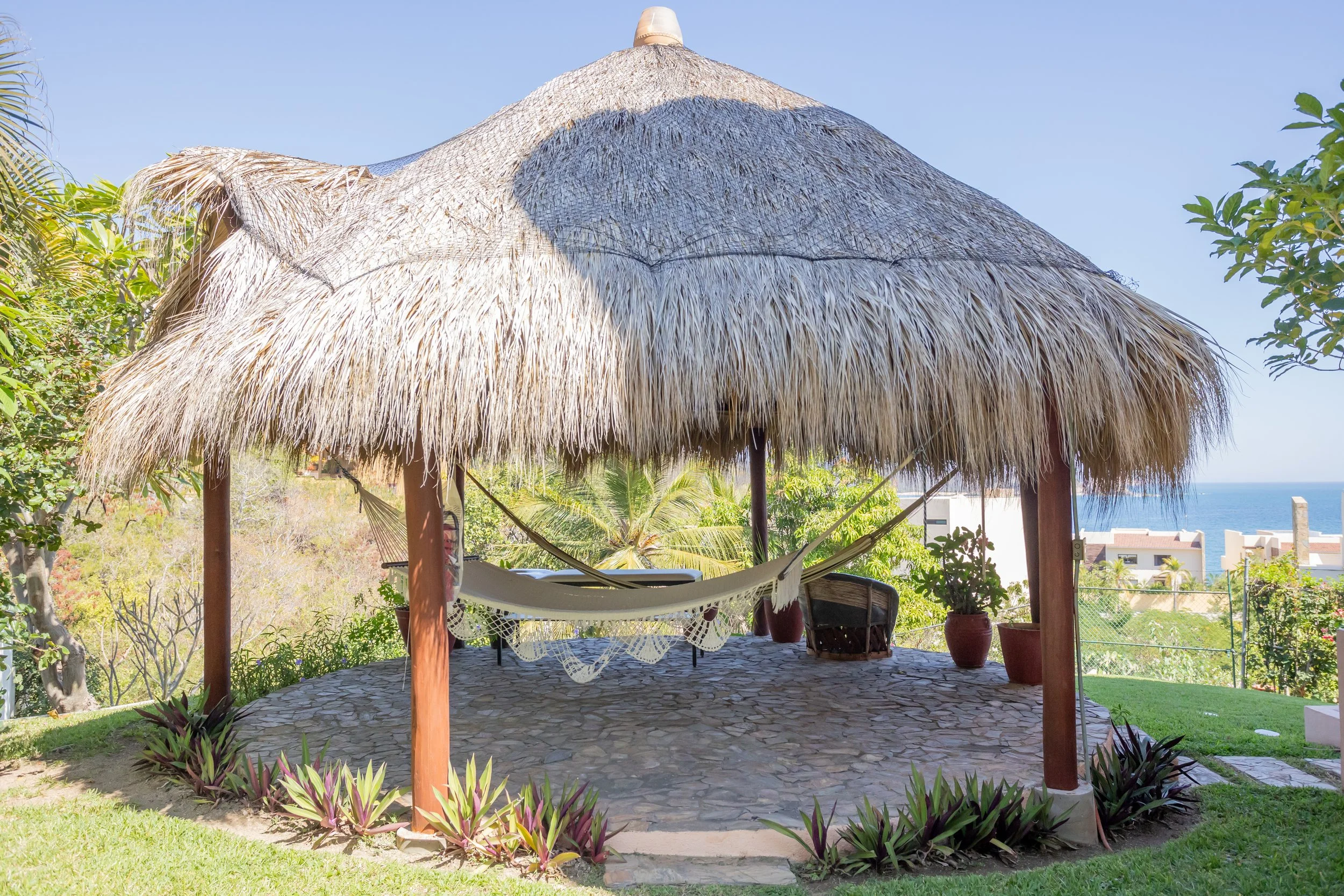 A thatched-roof palapa with a hammock underneath, surrounded by potted plants and tropical vegetation, overlooking the ocean and a nearby building in the background.
