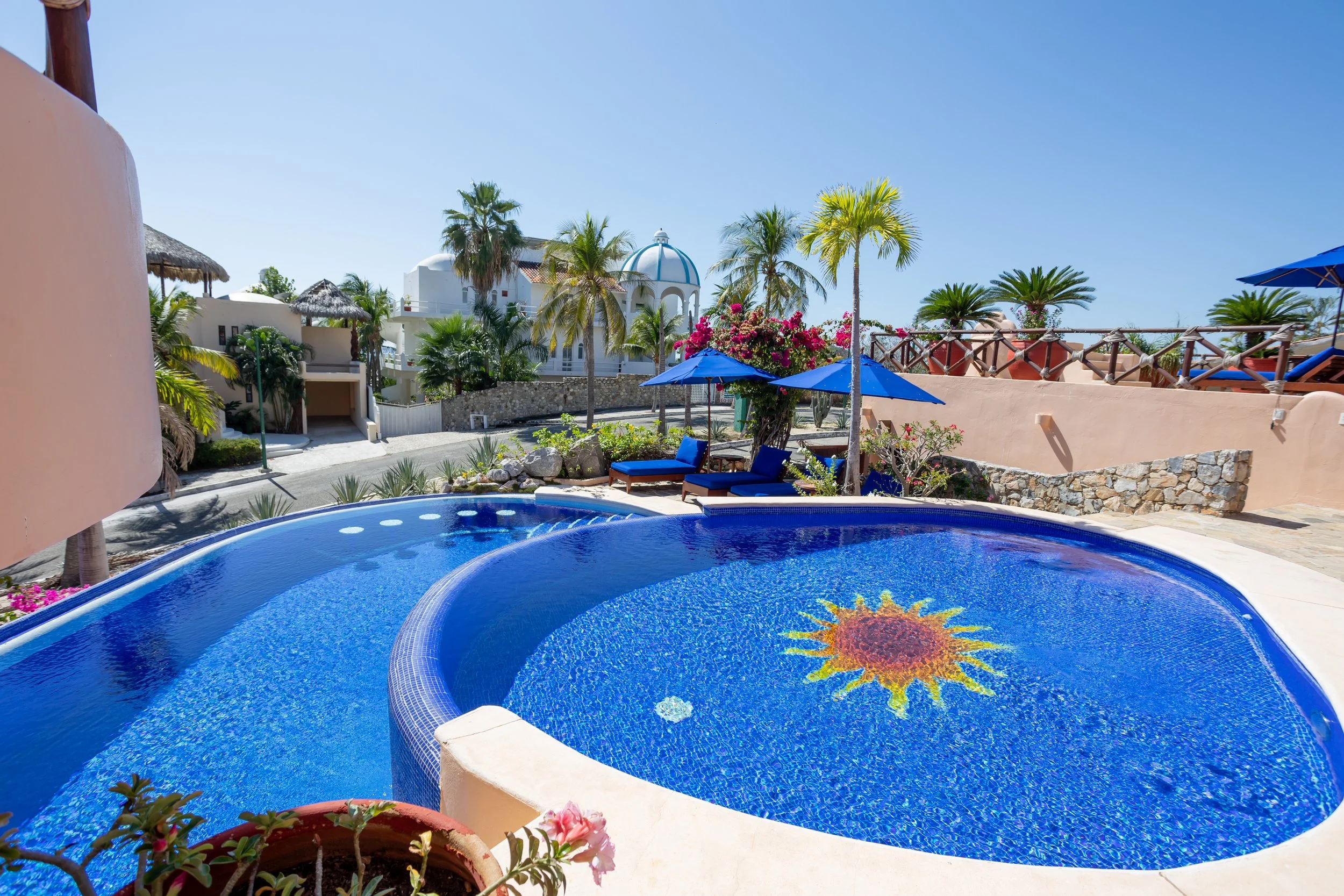 A view of a swimming pool with a colorful sun design at the bottom, surrounded by blue lounge chairs and umbrellas, palm trees, flowering bushes, and white buildings with a blue-domed structure in the background under a clear blue sky.