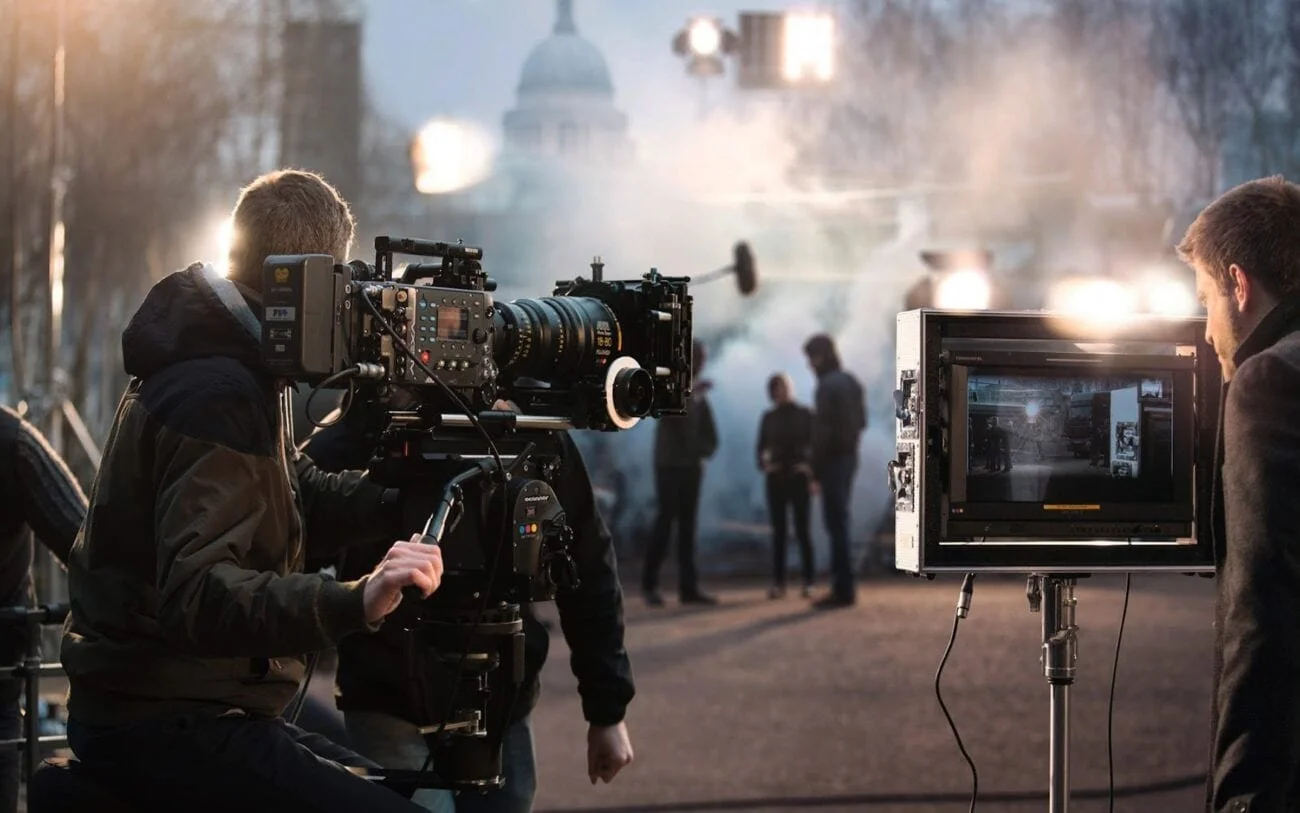 Filmmakers and camera crew shooting a scene outdoors with a large camera and monitor set-up, people in the background with blurred buildings and trees.