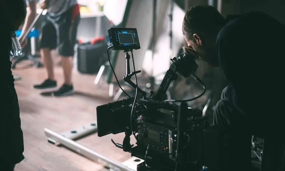 Filmmaker looking through a camera's viewfinder on set, with crew members in the background.