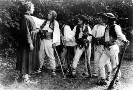 A woman in period clothing talking to a group of explorers or hikers in outdoor attire, with some holding walking sticks.