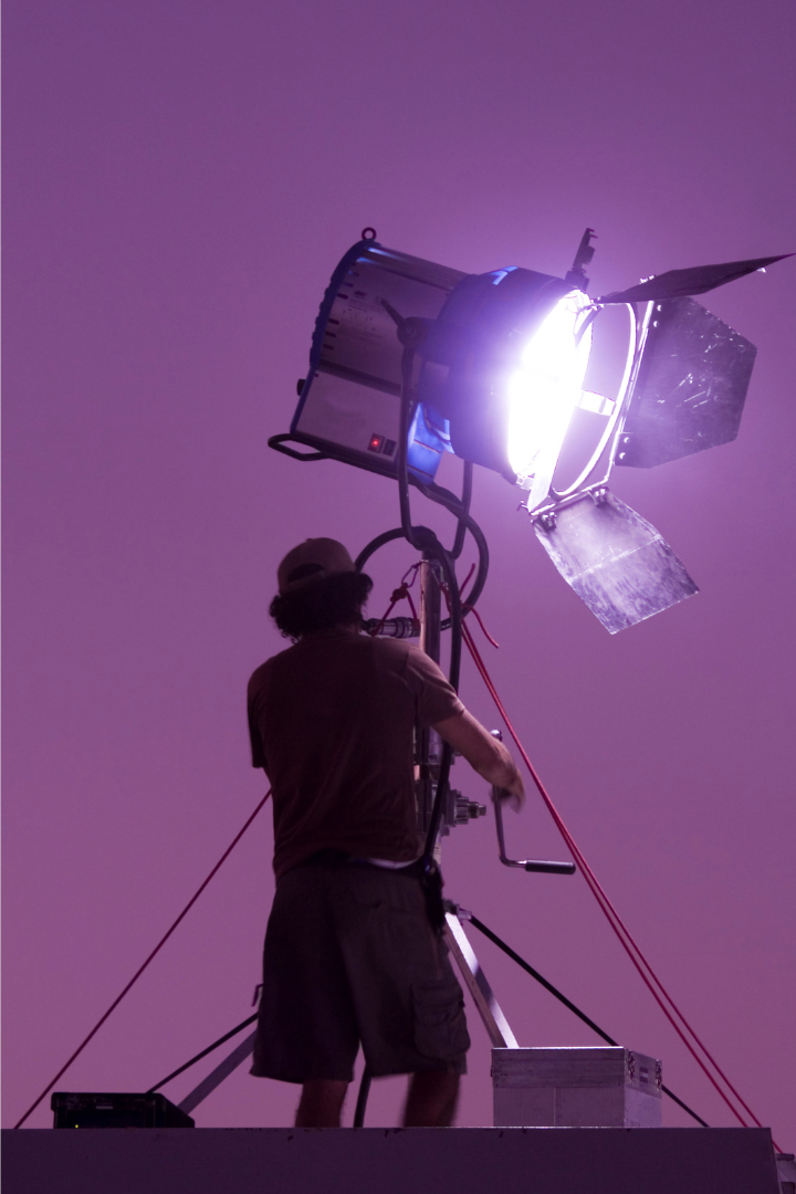 A person in casual clothing and a cap working on a large video or film lighting fixture against a purple background.