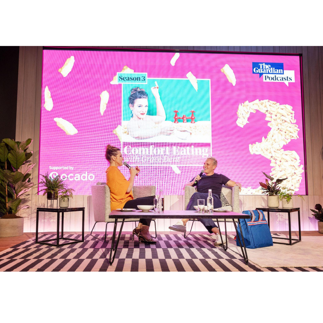 Two people are having a conversation on stage with a large pink screen behind them displaying promotional content for a podcast. The woman is wearing an orange dress and holding a microphone, while the man is in a black t-shirt and jeans, also holding a microphone. There are plants on either side of the stage, and a striped rug on the floor.