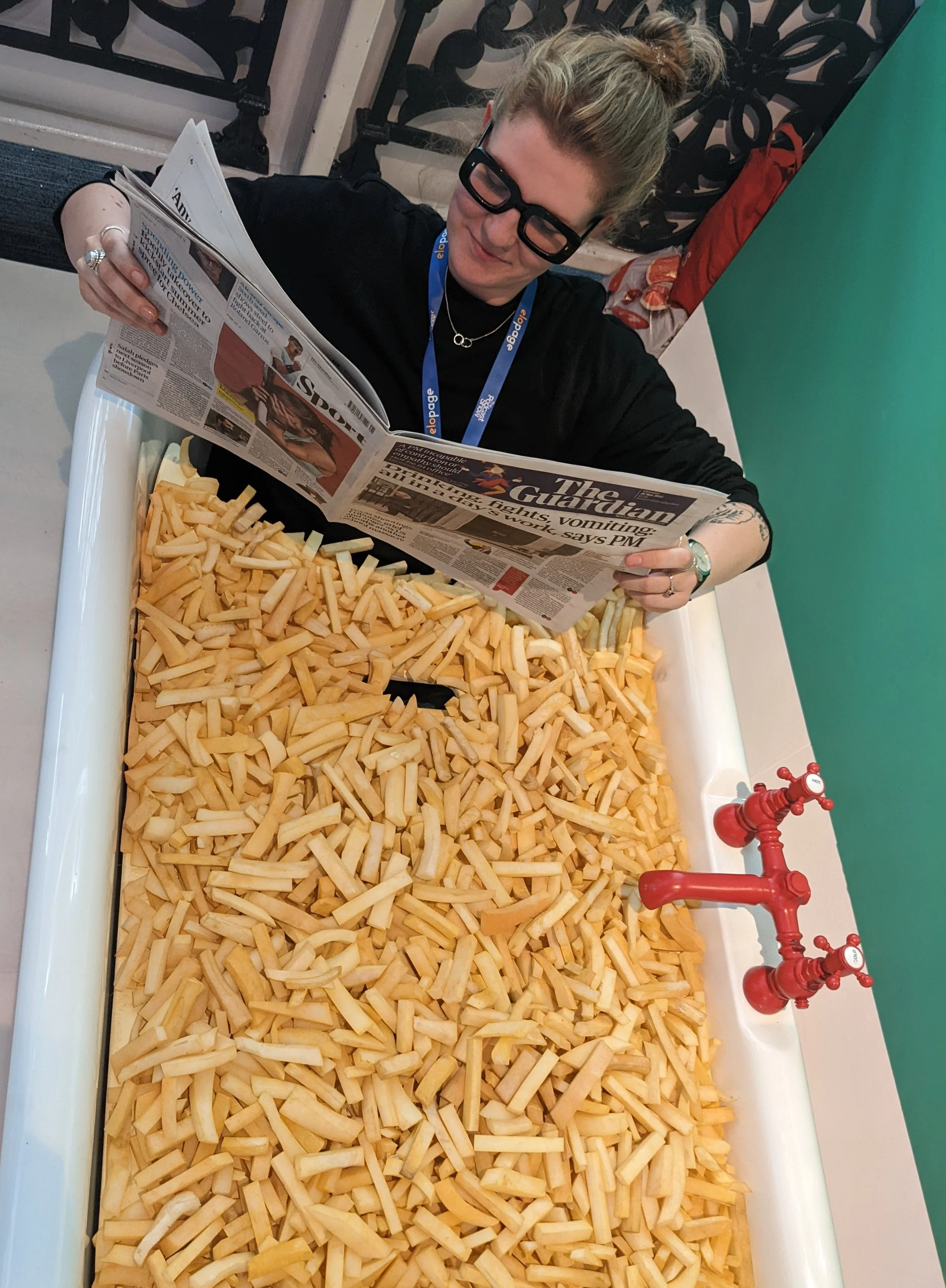 A woman wearing glasses reads a newspaper while sitting in a bathtub filled with french fries.