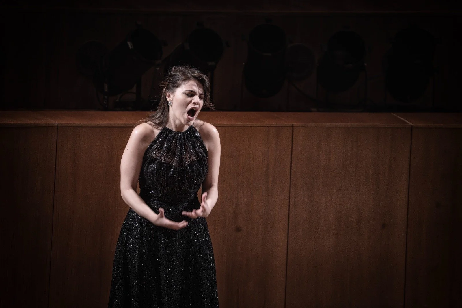 Una donna in abito nero si esibisce cantando con espressione intensa in un palco di un teatro o sala da concerto.