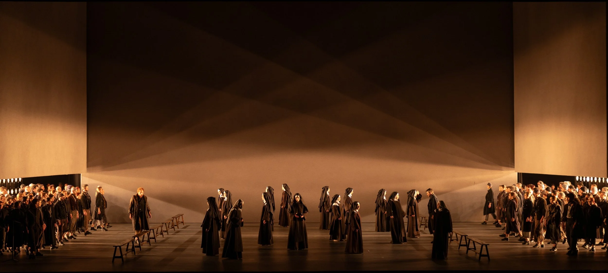 Spettacolo teatrale con molti attori vestiti con abiti scuri, in scena sotto una grande acquario nero con luci che creano ombre suggestive.