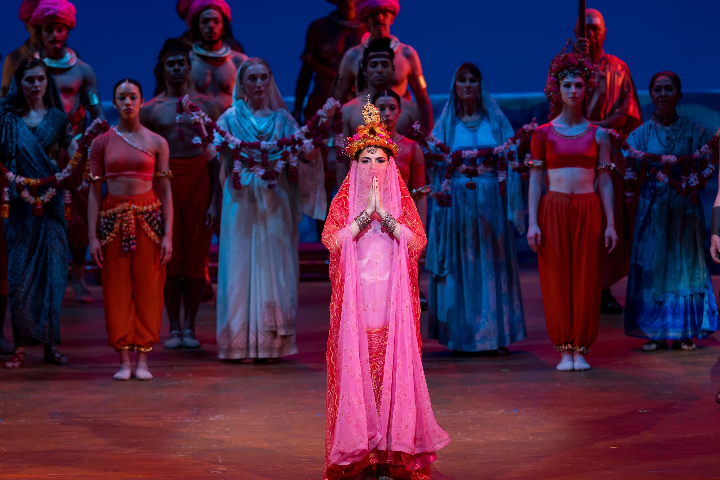 I Pescatori di Perle, Opera Palm Beach, Un gruppo di danzatori sul palco , con una donna al centro vestita con un abito rosa e un copricapo elaborato, mentre gli altri intorno indossano abiti colorati e decorati.