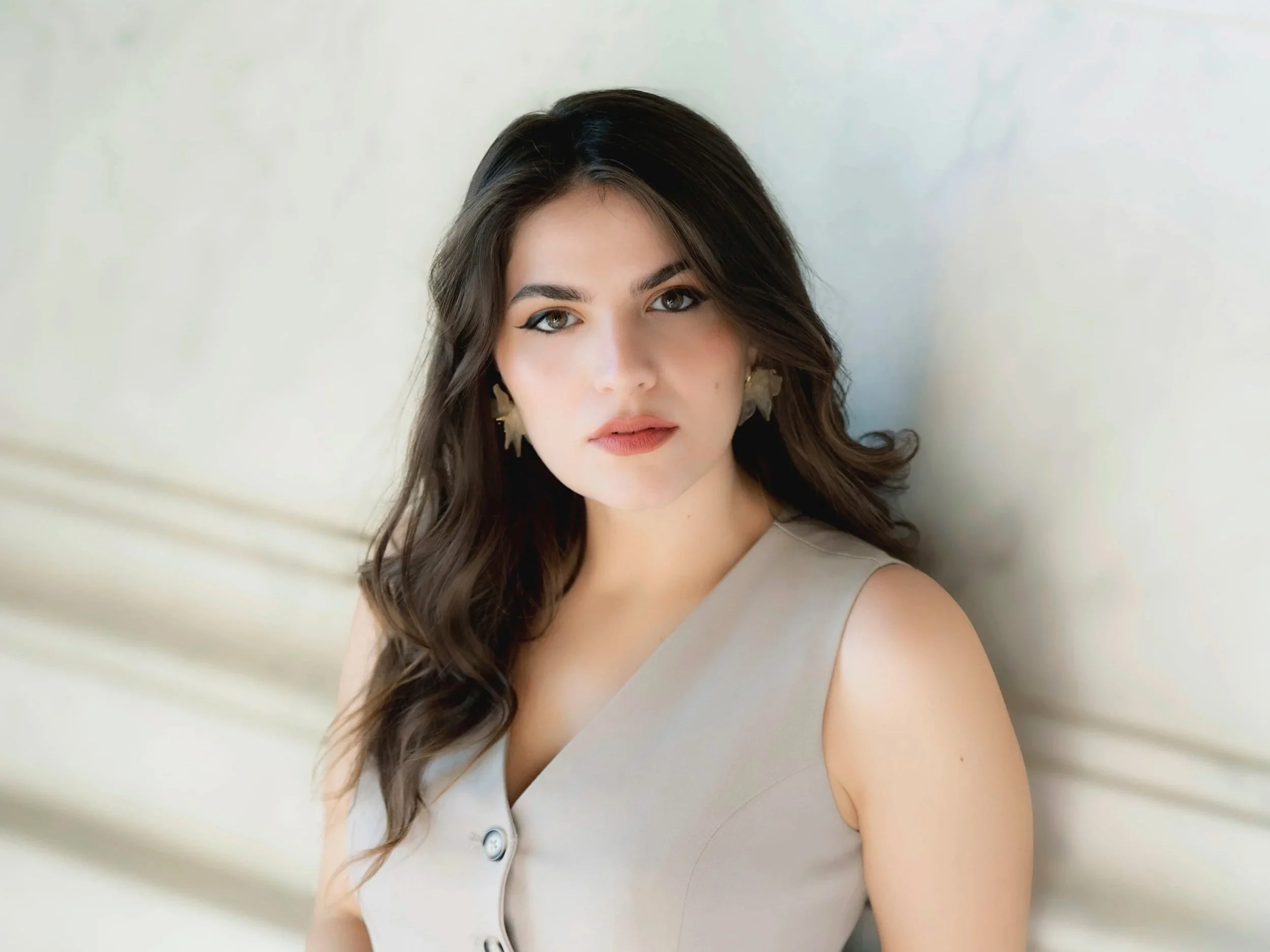 Francesca Pia Vitale, soprano con capelli castani ondulati, trucco naturale con eyeliner evidente, indossa orecchini a forma di foglia e un vestito senza spalline di colore grigio chiaro, posa davanti a una parete bianca.