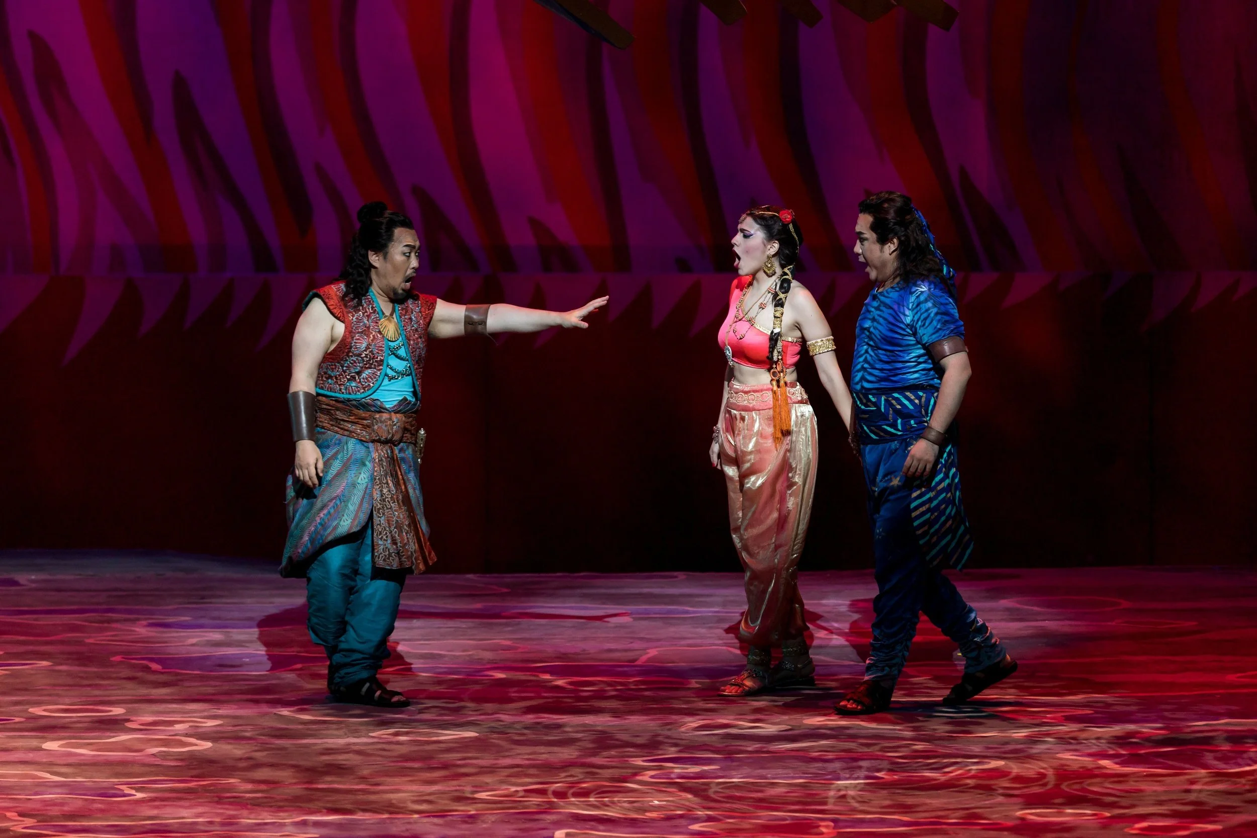 Scena teatrale con tre attori in costume colorato, due uomini e una donna, durante una performance sul palco con sfondo viola e rosso.