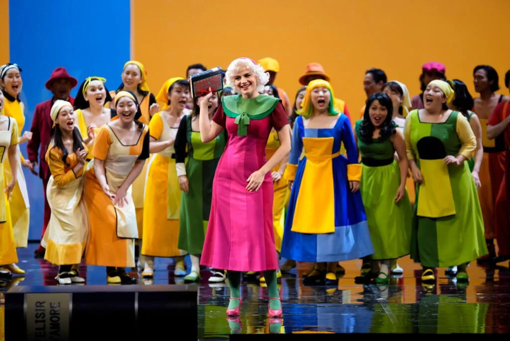Una scena di teatro con molte persone vestite con abiti colorati e un personaggio in primo piano con capelli bianchi, vestito rosa e verde, che tiene un tablet.