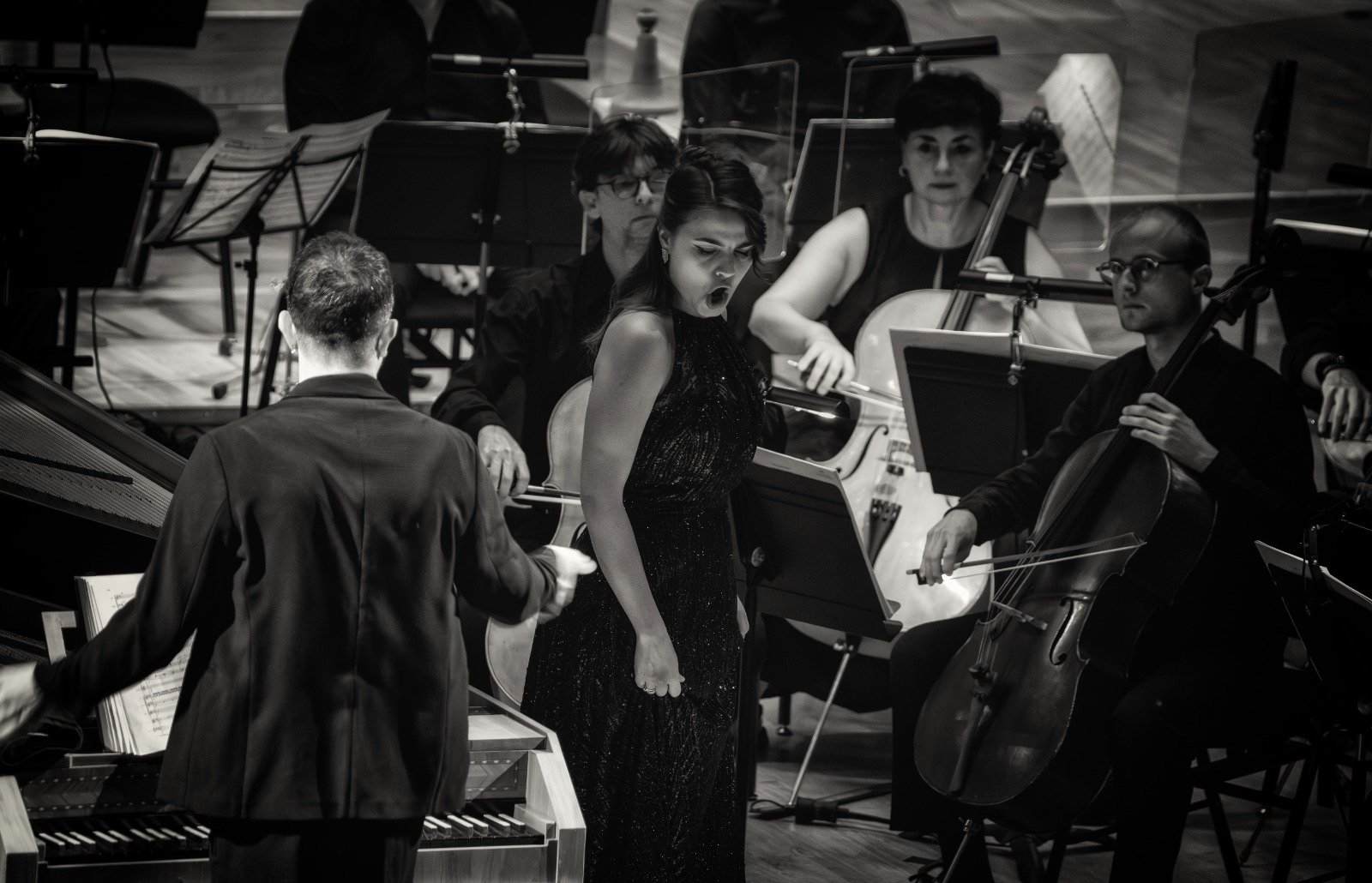 Un'orchestra al lavoro con un direttore, un'triodenne donna canta con passione, e altri musicisti con strumenti come violoncello, violino, e pianoforte, in una sala da concerto.