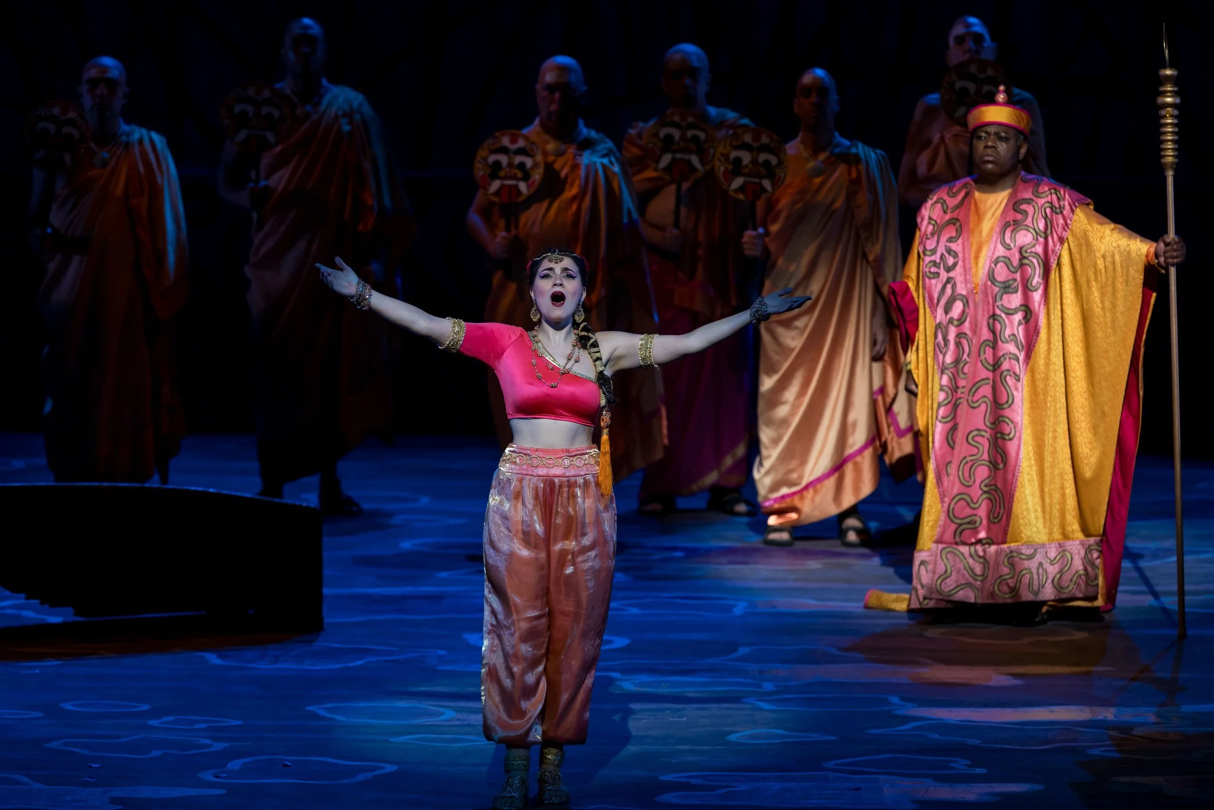 Una scena di un'opera teatrale con un'attrice al centro che canta, vestita con abiti tradizionali rosa e oro, circondata da uomini con maschere e abiti di scena sulle spalle in un teatro.