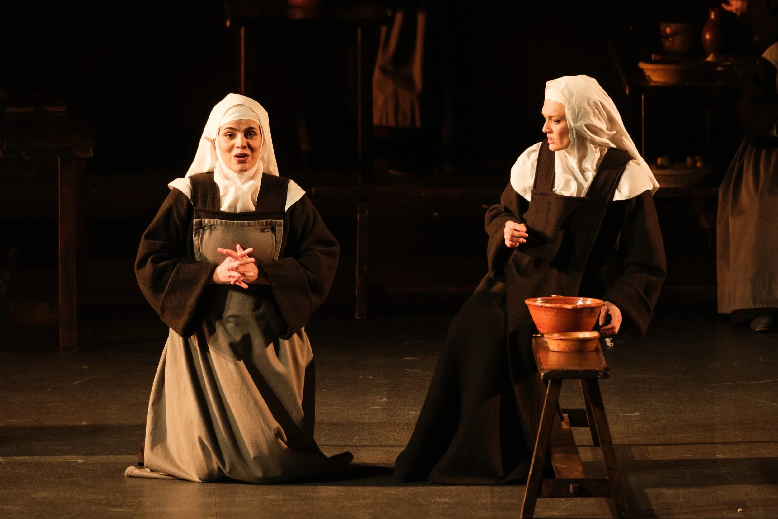 Due attrici in costumi d'epoca sul palco durante una rappresentazione teatrale, una di fronte e l'altra di fianco, con scene di scena in background e due recipienti di terracotta su una sedia.