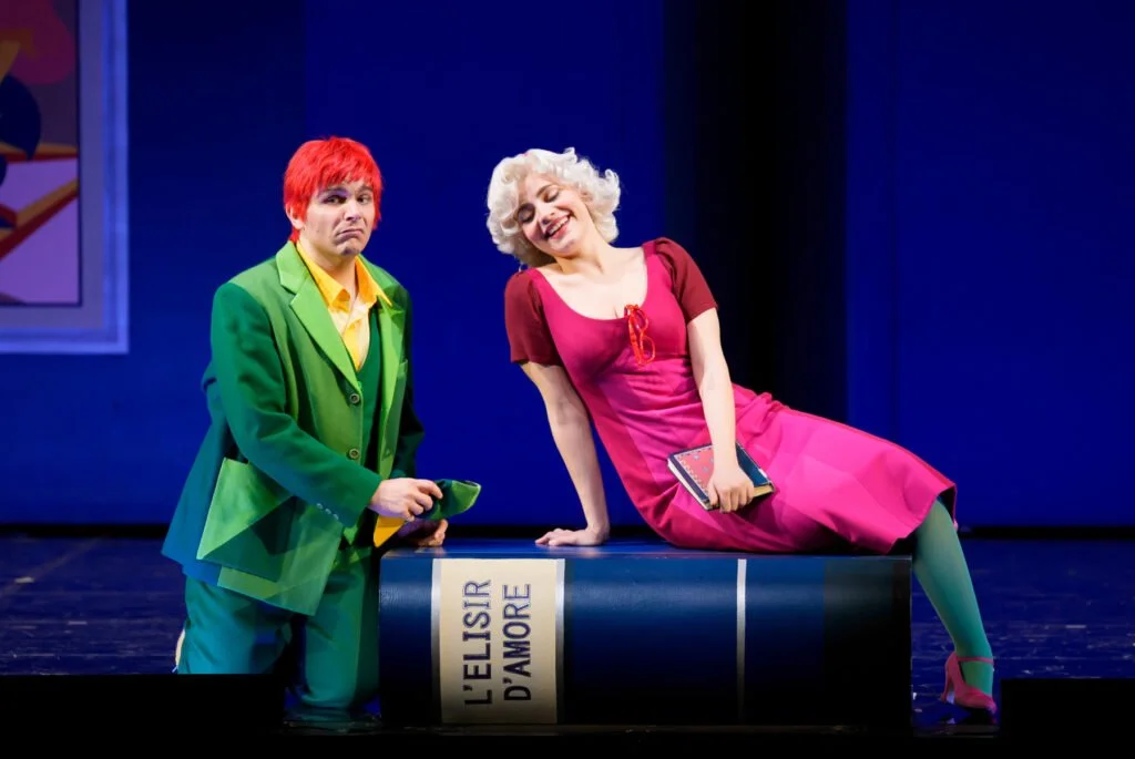 Due attori travestiti interpretano una scena teatrale, uno con capelli rossi e abiti verdi, l'altra con capelli bianchi e abiti rosa, in un teatro con sfondo blu