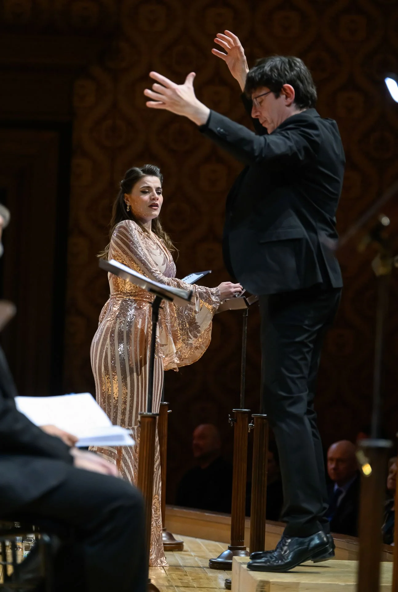 Un uomo dirige un'orchestra, mentre una donna in abito elegante canta durante un'esibizione in un teatro.
