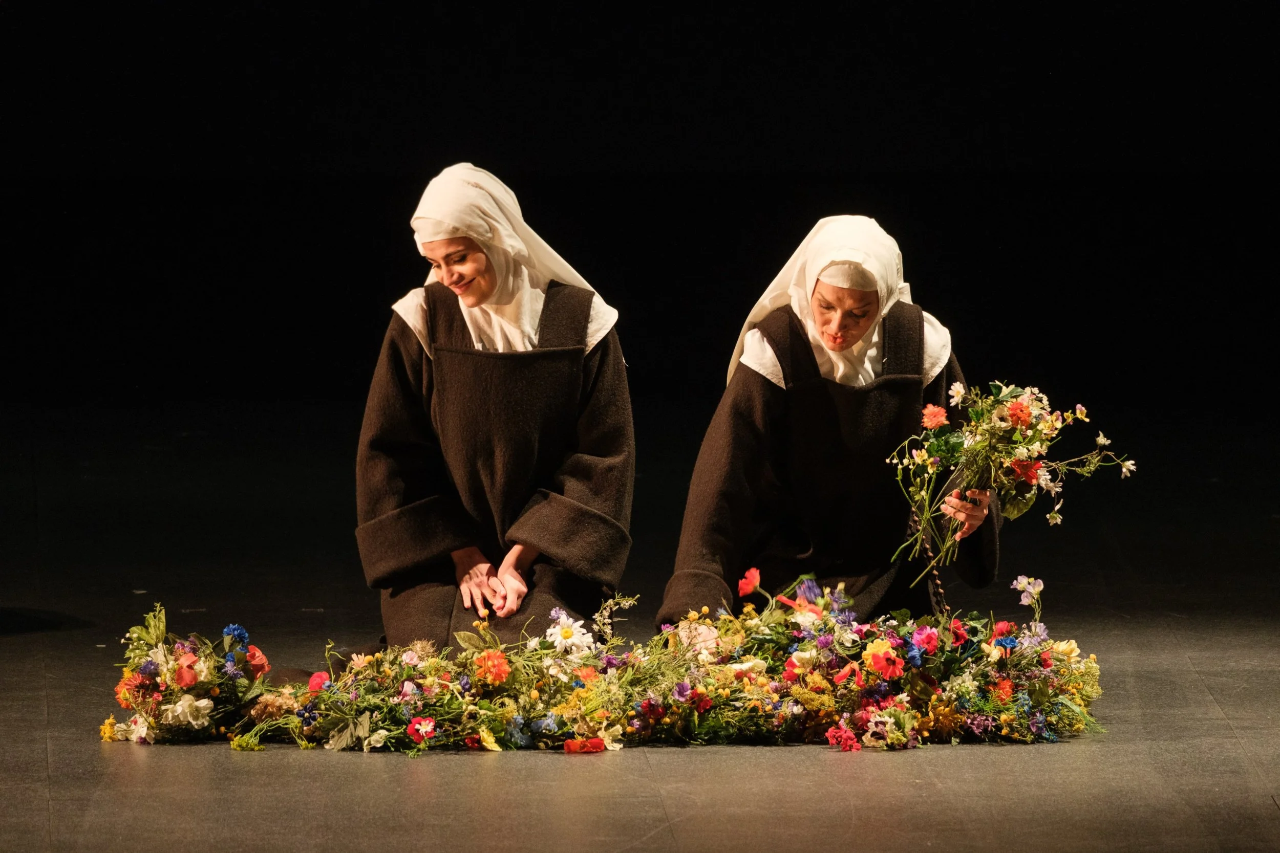 Due donne vestite con abiti neri e veli bianchi sul capo, piegate in avanti mentre sistemano un tappeto di fiori colorati sul palco, in un ambiente scuro e senza pubblico visibile.