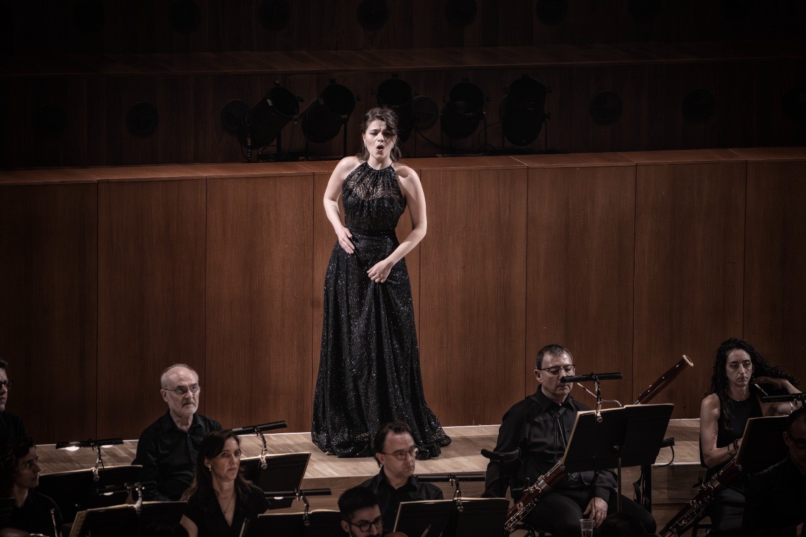 Una donna in abito lungo nero mentre canta durante un concerto con un'orchestra, sul palco in un teatro