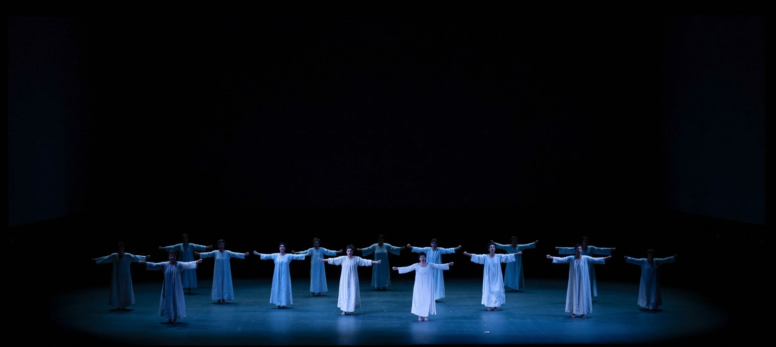 Gruppo di persone, probabilmente attori, in costume bianco, che si trova su un palco con uno sfondo nero, durante una performance teatrale o di danza