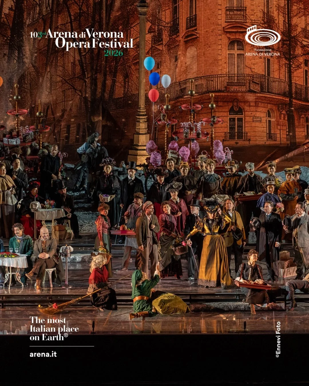 Scena di un'opera teatrale ambientata a Verona, con personaggi in costumi d'epoca sul palcoscenico. Sullo sfondo, un edificio storico e decorazioni teatrali colorate riconducibili al Festival dell'Opera di Arena di Verona 2026.