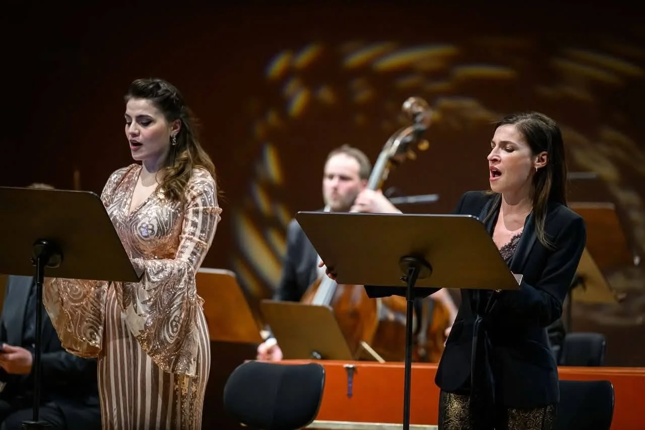 Due donne cantano durante un concerto, con un musicista che suona il contrabbasso sullo sfondo. Entrambe sono di profilo, con abiti eleganti e leggins lucidi, mentre si esibiscono davanti a supporti per musica.