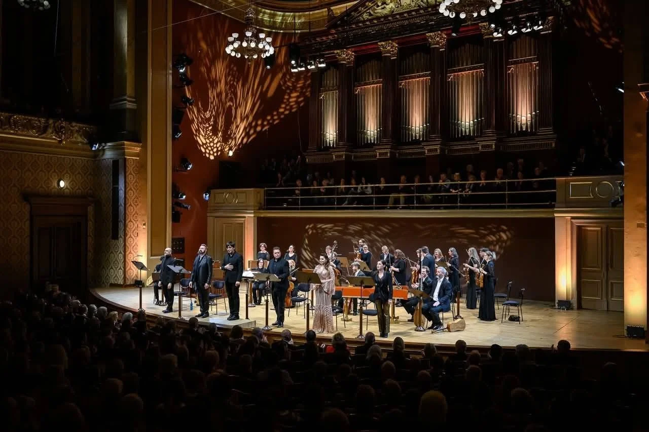 Un'orchestra sul palco di un teatro, composta da musicisti con strumenti musicali, con pubblico seduto che guarda. L'ambiente è elegante con illuminazione calda e tendaggi sullo sfondo.