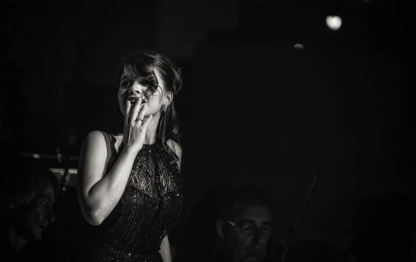 Donna in abito elegante con capelli mossi, in un ambiente di festa o concerto, con altre persone sullo sfondo.