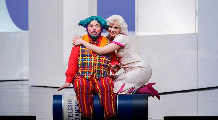 Due attori in costumi teatrali, un uomo con parrucca blu e abiti colorati seduto su una scatola, una donna con capelli bianchi e vestito rosa che lo abbraccia sorridendo.