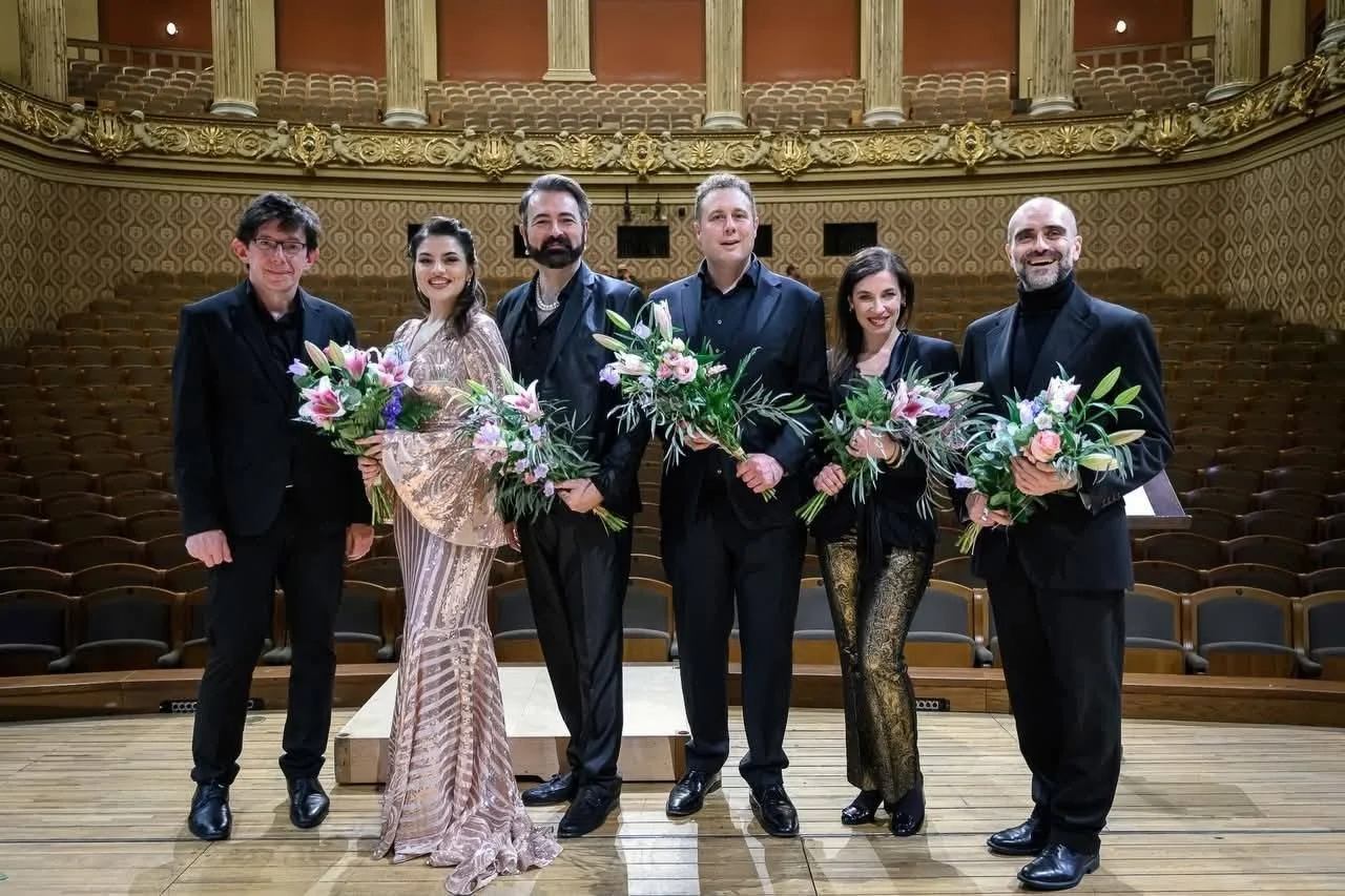 Gruppo di sei persone in abiti eleganti, con alcuni che tengono bouquet di fiori, sul palco di un teatro con decorazioni dorate e sedie vuote alle spalle.