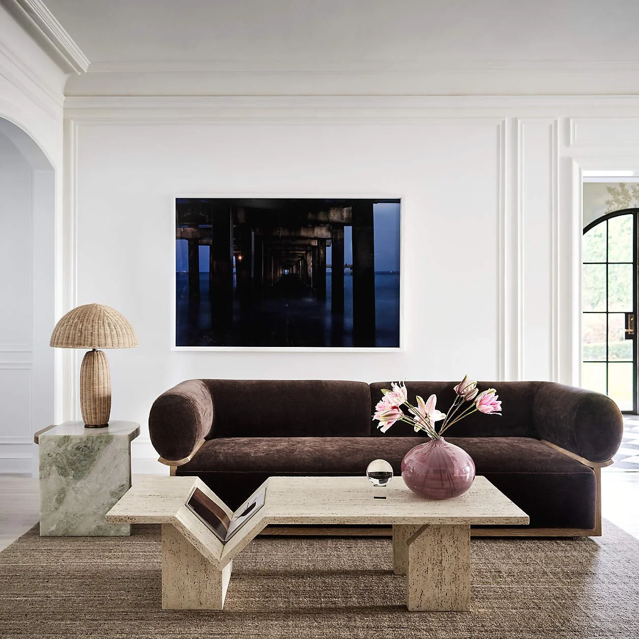 Living room with a dark brown velvet sofa, a marble side table with a wicker lamp, and a rectangular marble coffee table with a pink vase of lilies and a small glass orb. A large framed photo of an ocean pier at night hangs on the white wall behind the sofa, with a window to the right.