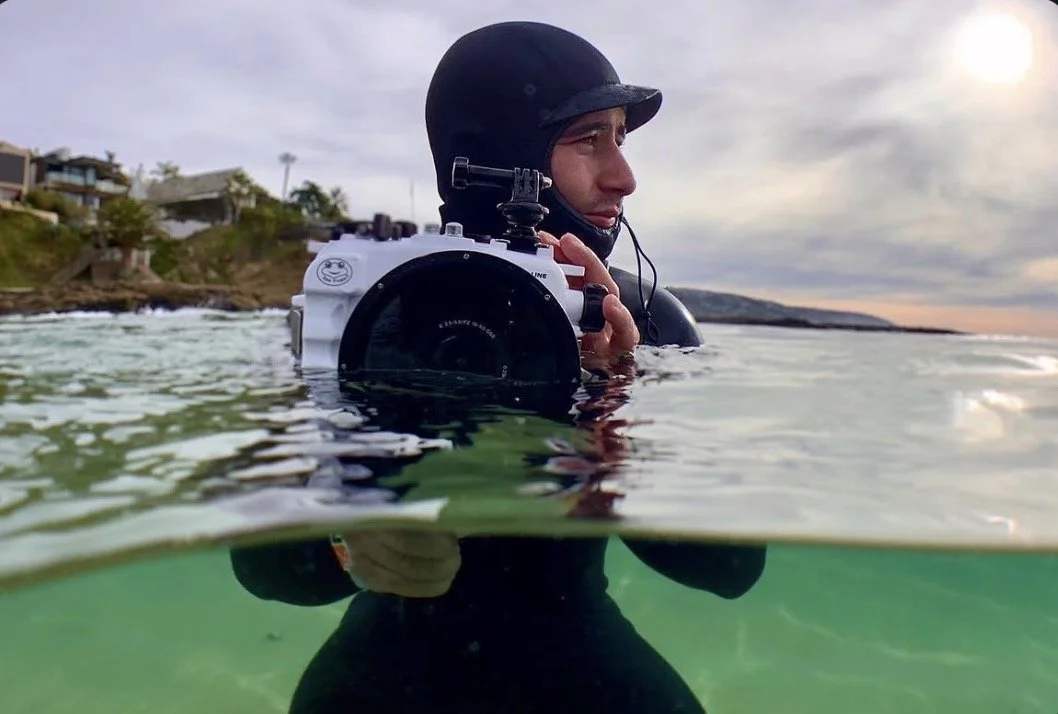 A diver wearing a black helmet and wetsuit, holding a camera while floating in the water with a coastal landscape in the background.
