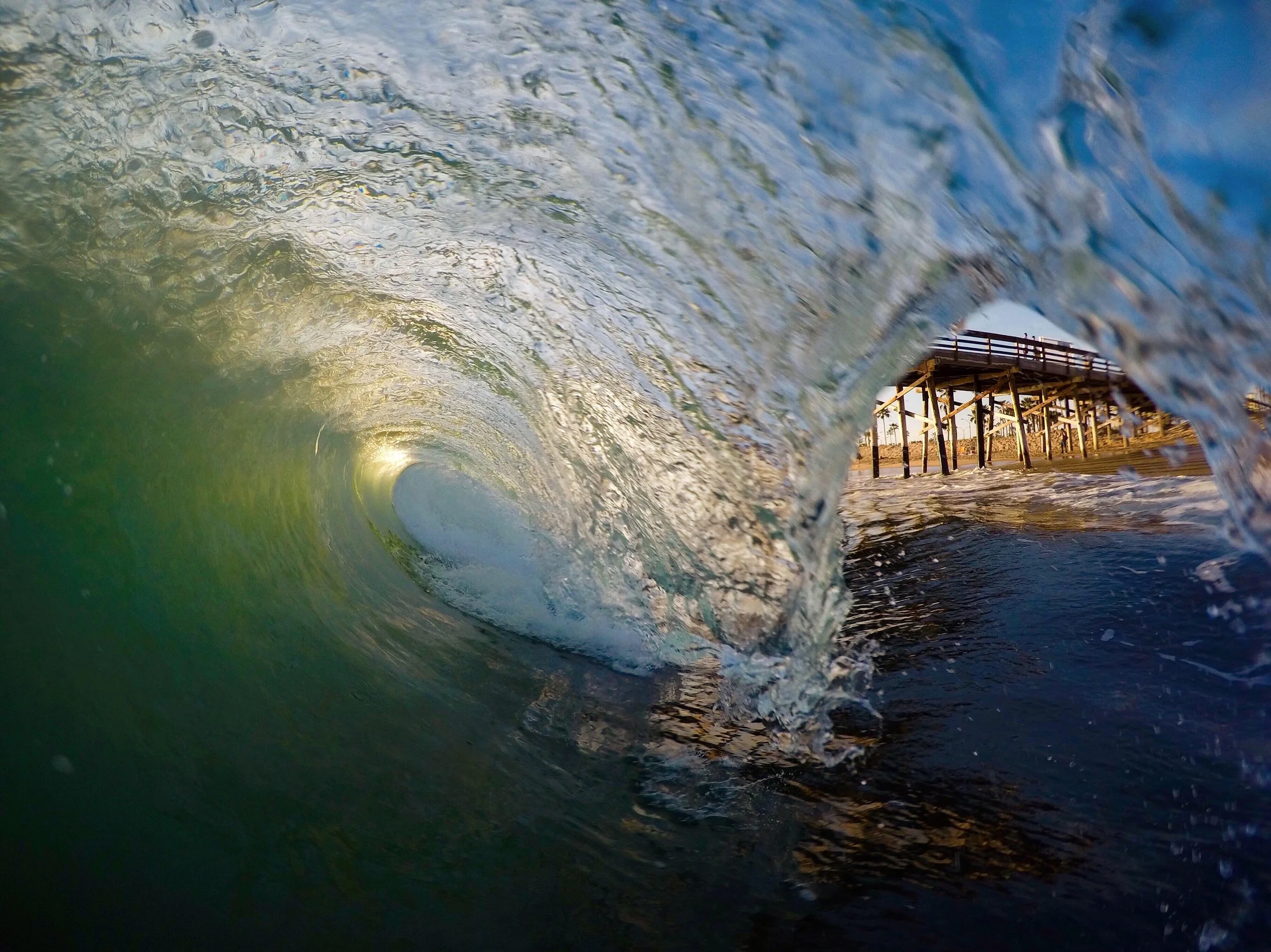 View from inside a breaking ocean wave, with a wooden pier and sunset in the background.