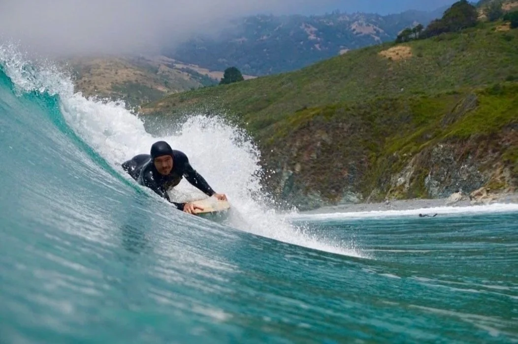 Person surfing on a wave near a green hillside during daytime.