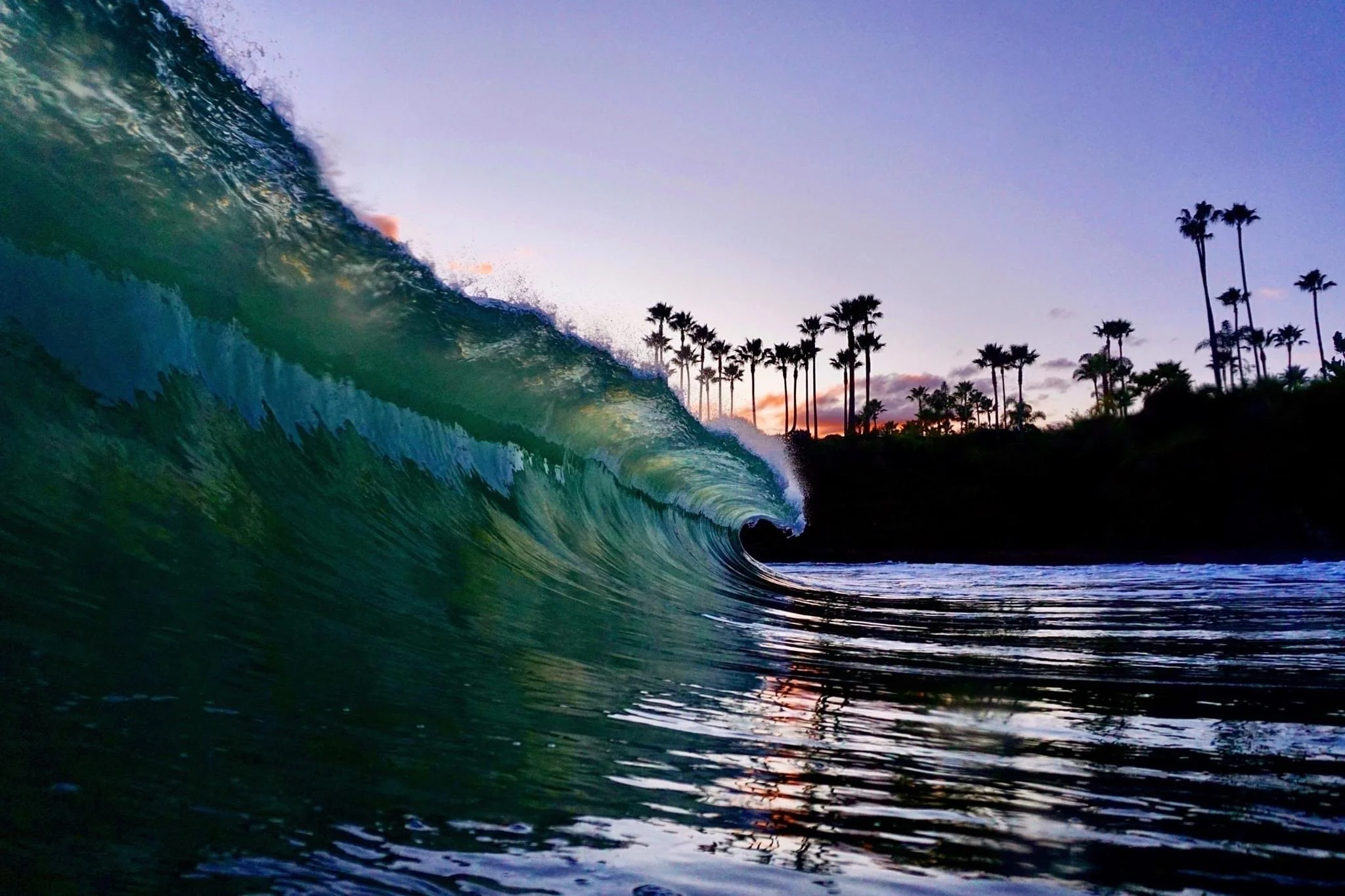 Surf breaking on a beach at sunset with palm trees in the background.