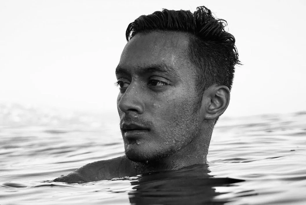 A young man with wet hair and water droplets on his face, partially submerged in water, looking off to the side in black and white.