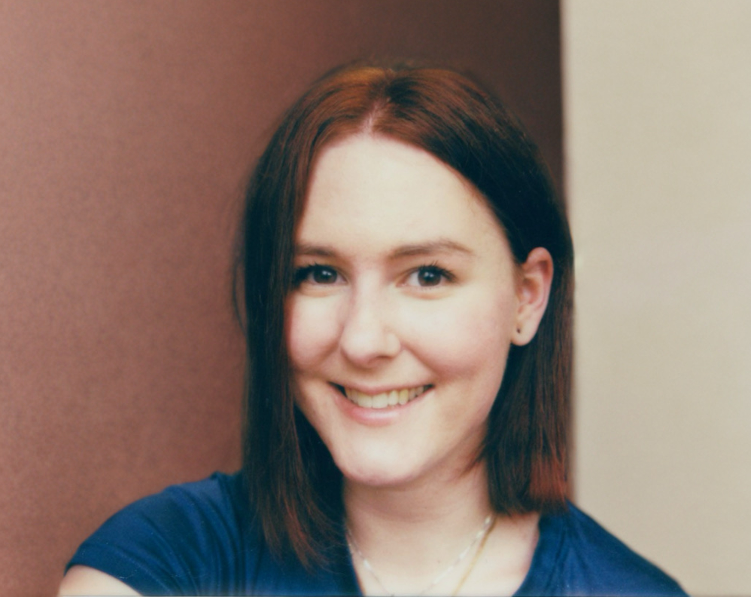 Close-up of a woman with dark brown hair, smiling, wearing a blue shirt, standing against a multi-colored background.