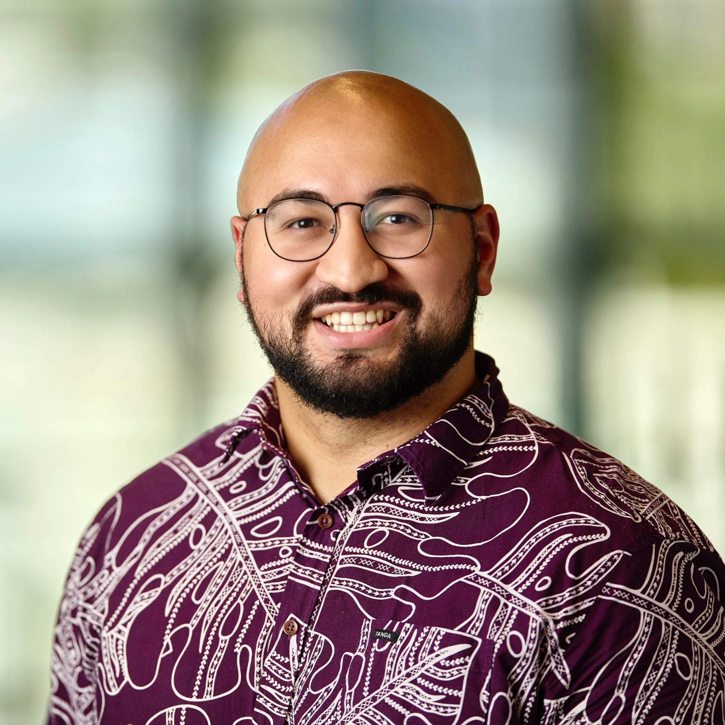 Dr. Ocean Akaka smiling, wearing a purple shirt with white patterns, standing against a blurred green background.