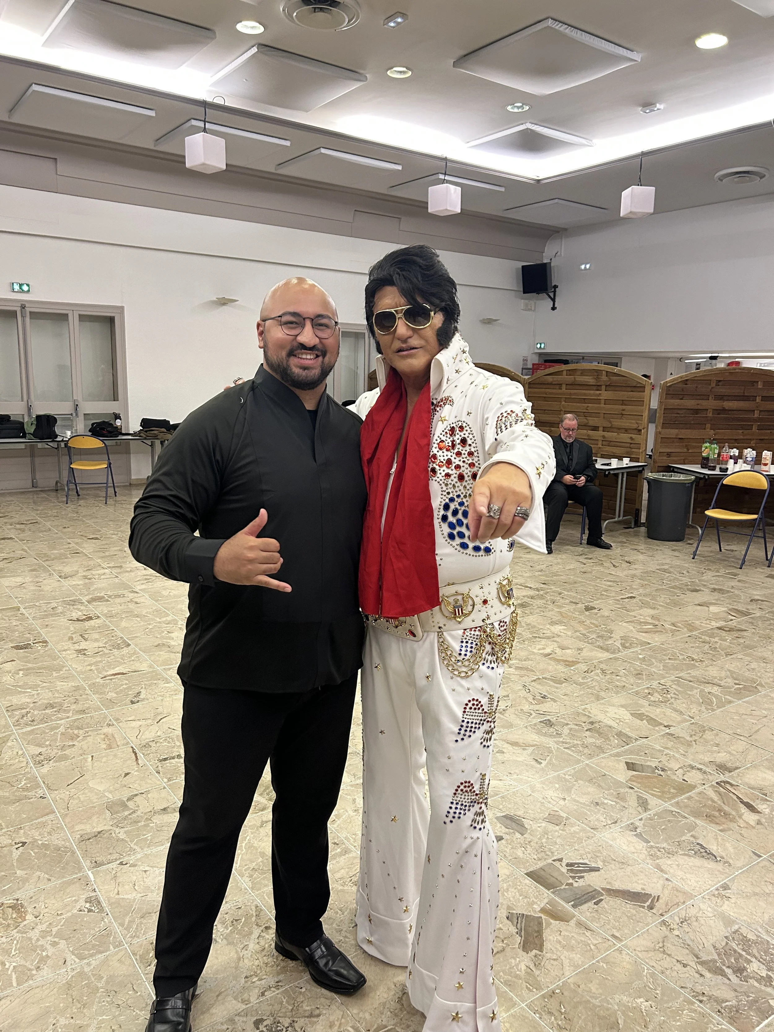 Dr. Akaka dressed in black and a performer dressed as Elvis Presley, wearing sunglasses, a sequined jumpsuit with stars and embellishments, posing together for a photo in a banquet hall with beige tile flooring and a decorated ceiling.