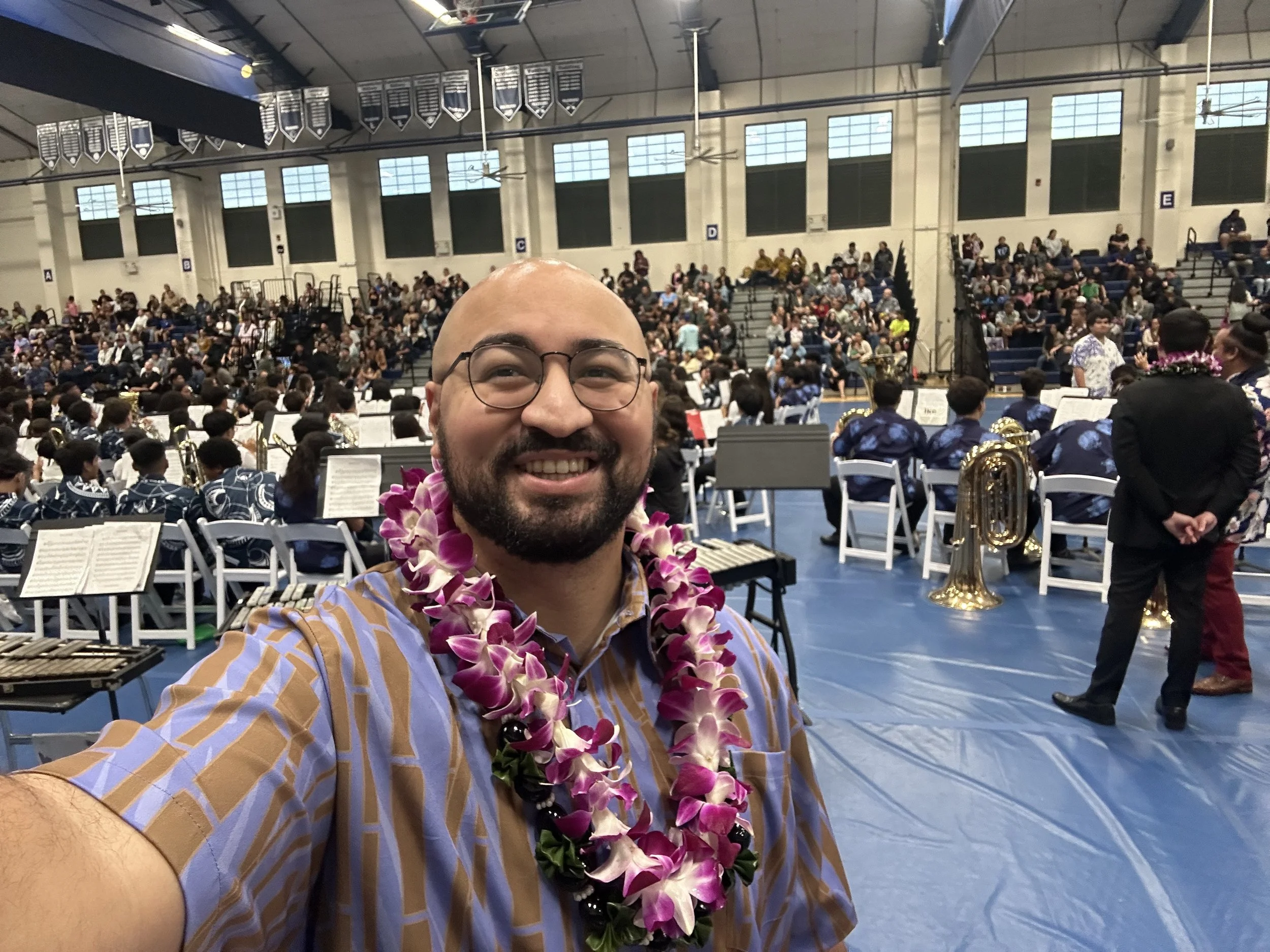 Dr. Ocean Akaka smiling in a gymnasium filled with seated audience and musicians in uniform, wearing a lei, during a concert.