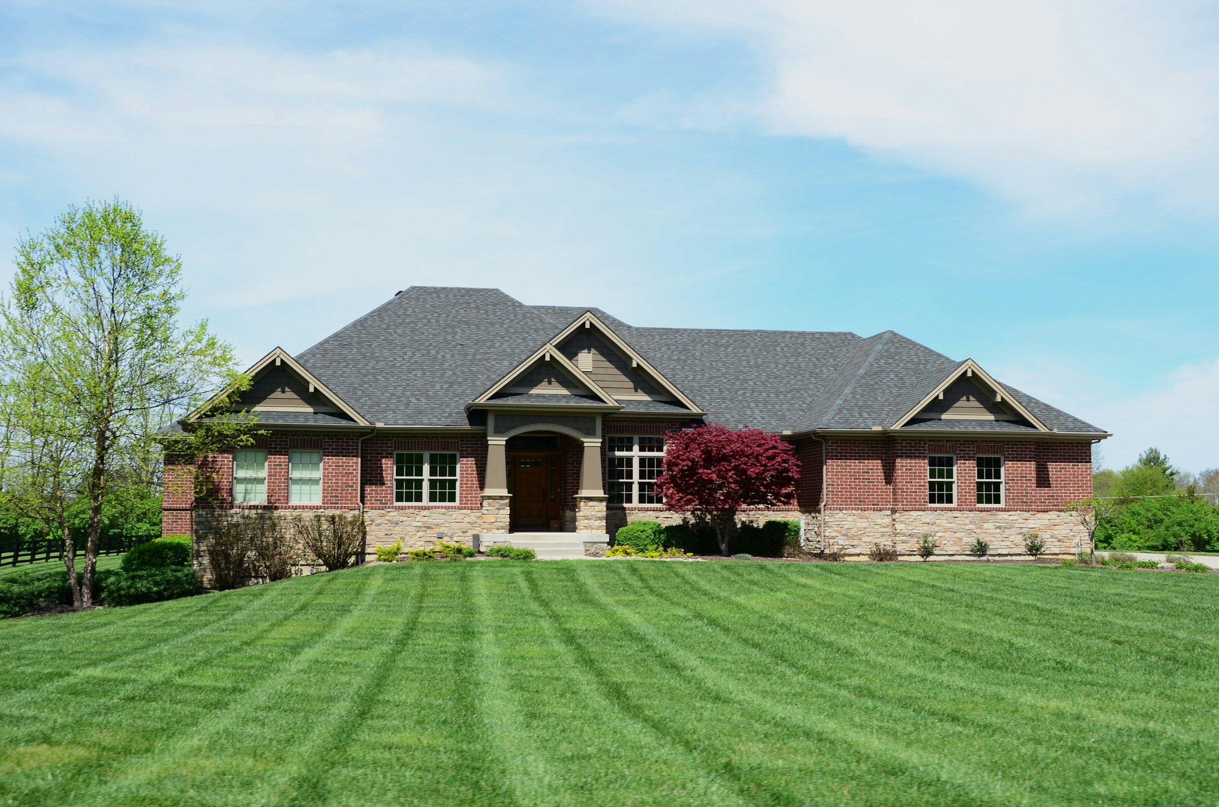 A large brick house with a gray shingle roof, surrounded by a well-maintained lawn and trees, under a partly cloudy sky.