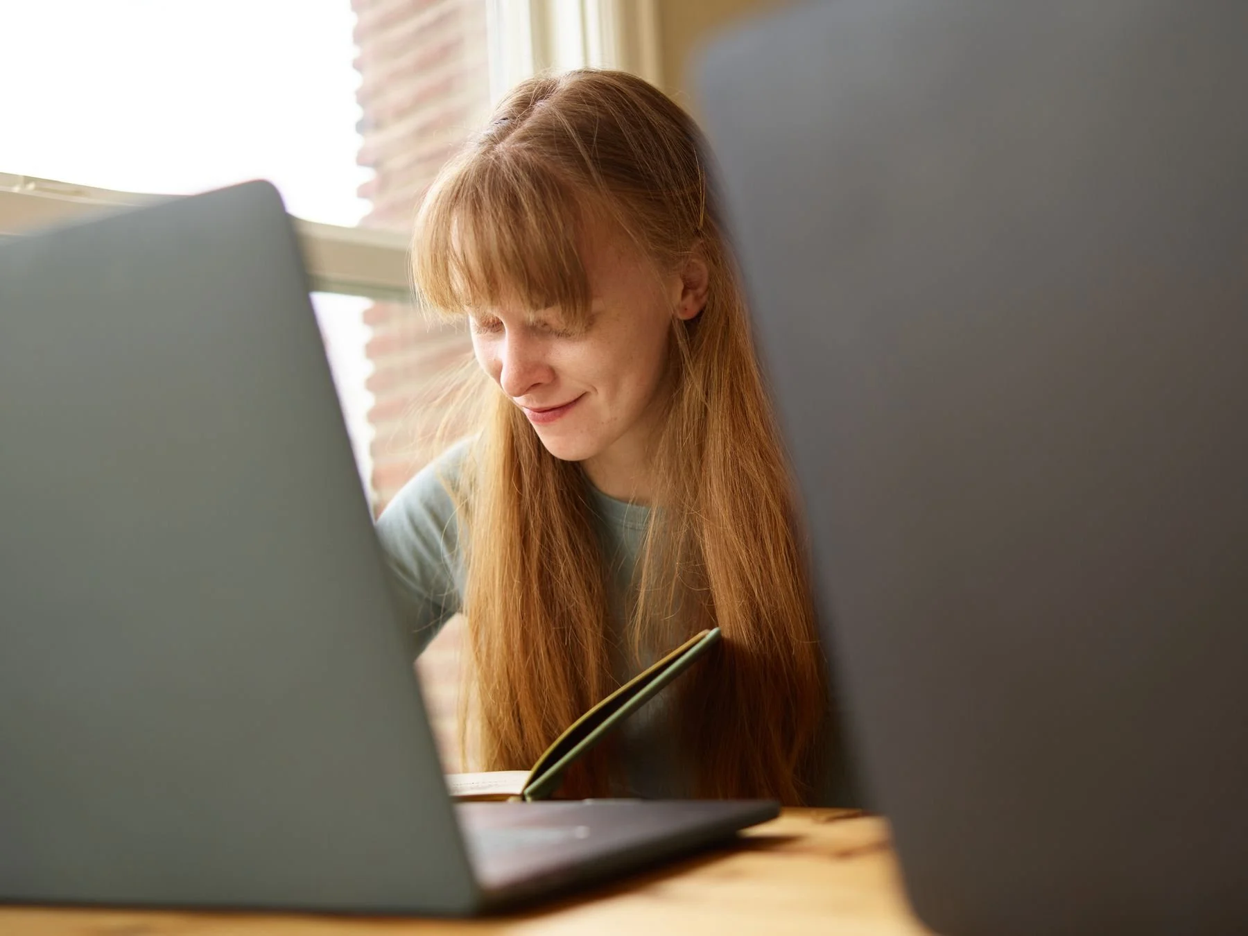 Woman using a laptop and notebook beside a sunlit window.
