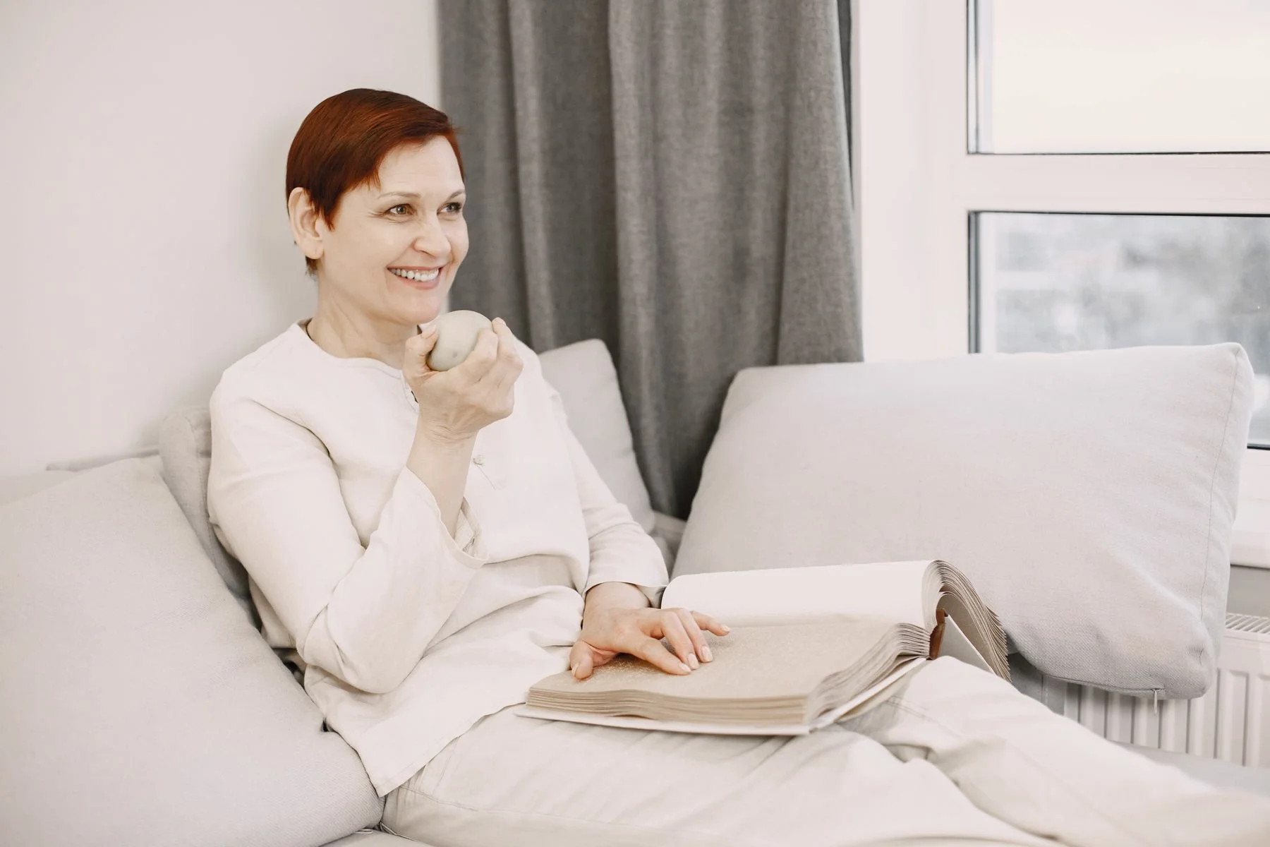 Woman relaxing with a book on a sofa beside a window.