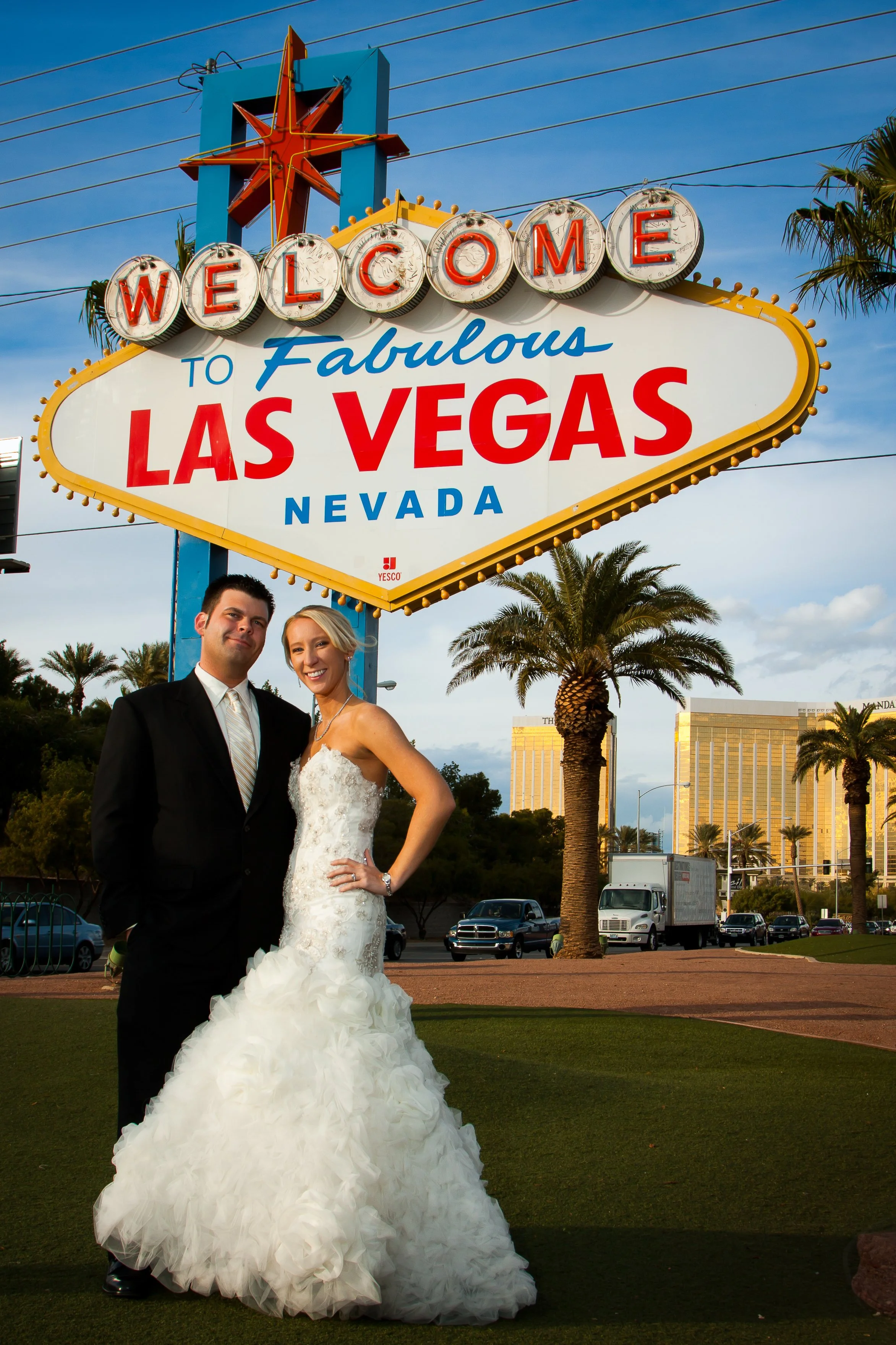 A newlywed couple posing in front of the Welcome to Fabulous Las Vegas sign, with palm trees and a cityscape in the background on a clear day.