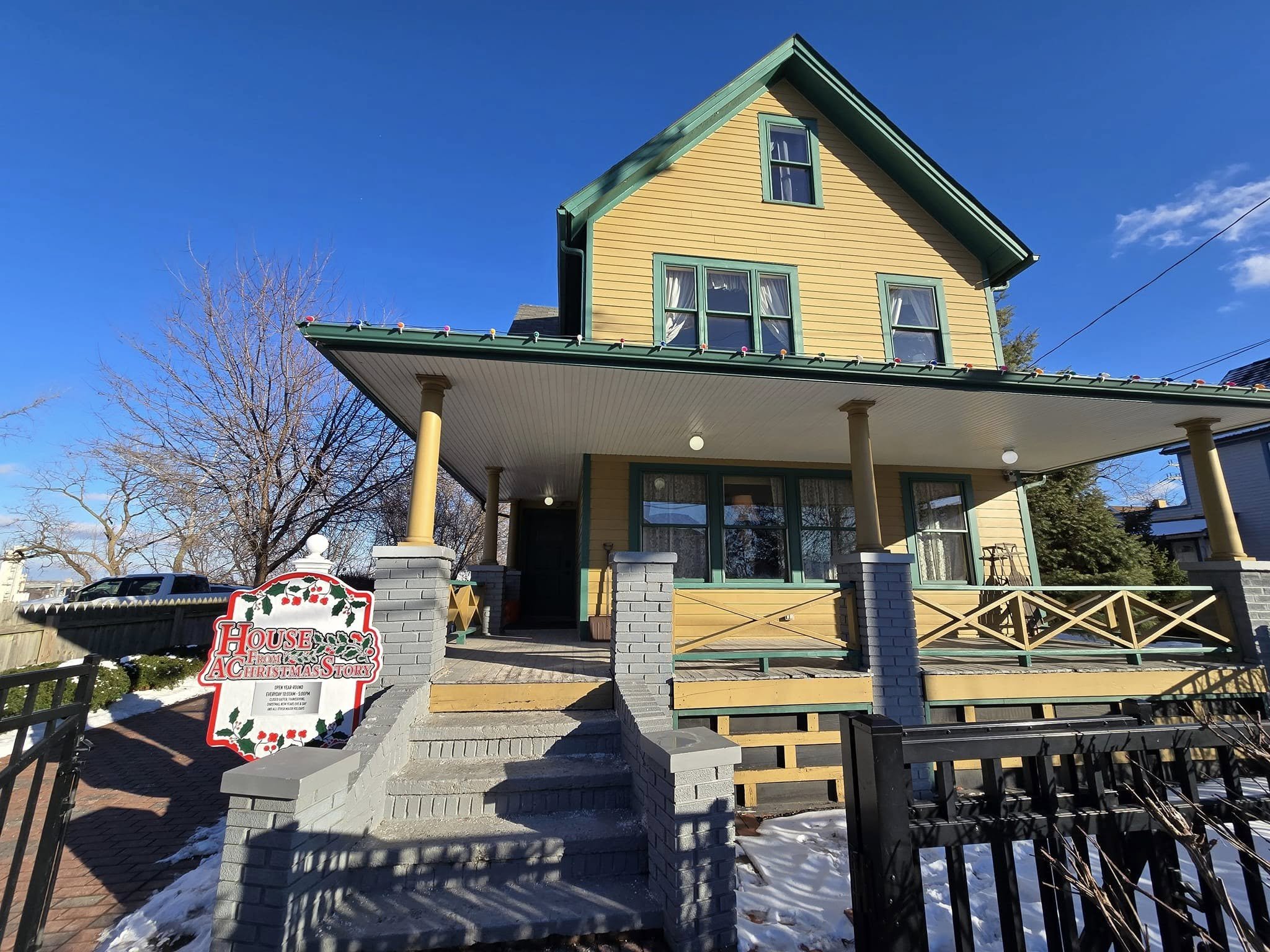 A yellow house with green trim and a porch, decorated with Christmas lights, and a sign that reads 'House from A Christmas Story' in front of the house on a paved path, with snow on the ground and bare trees around.